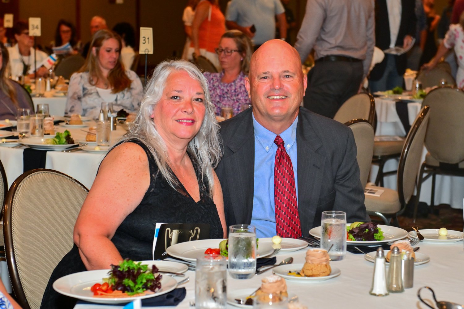 A middle-aged woman with long gray hair and a man with a bald head sit together at a banquet table during a formal event. The table is set with plates of salad, rolls, and glasses of water. Other guests are visible in the background, seated or walkin