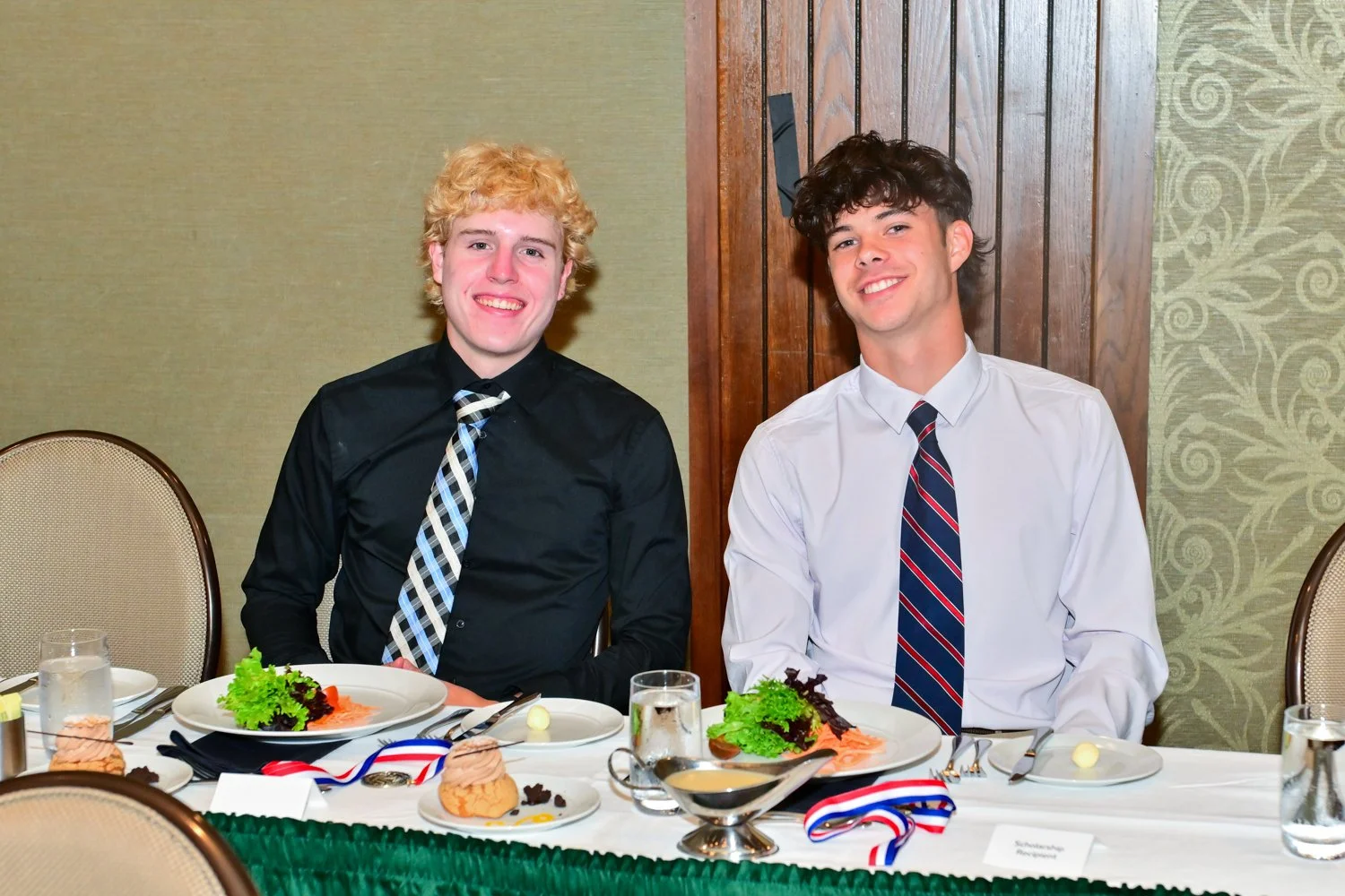 Two young men dressed in formal attire, sitting at a decorated banquet table with plates of food, medals, and water glasses, smiling at the camera in a banquet hall.