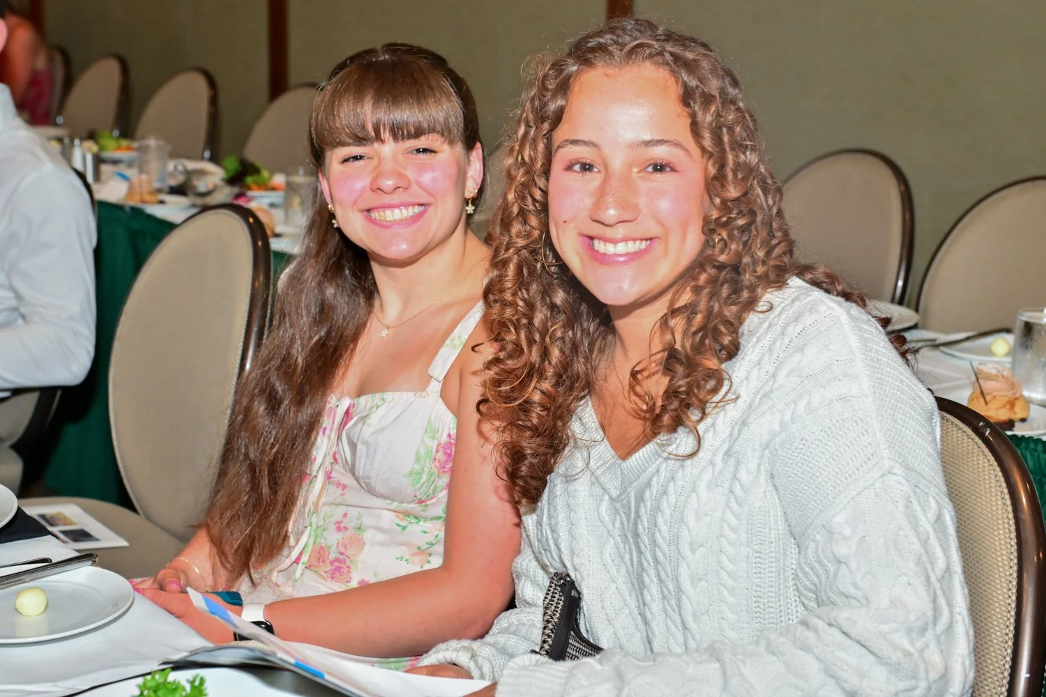 Two women sitting at a table, smiling at the camera at a social gathering or event, with plates and food on the table.
