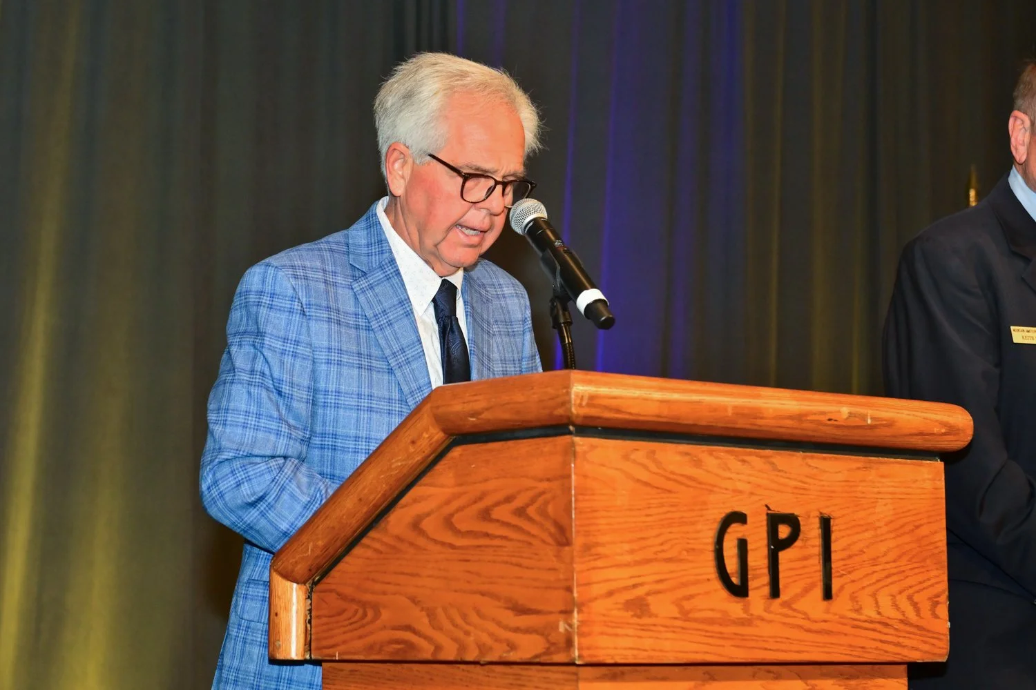 A man with glasses and gray hair, wearing a blue checkered suit and white shirt, is speaking at a wooden podium with the letters 'GPI' on it, in front of a dark curtain background.