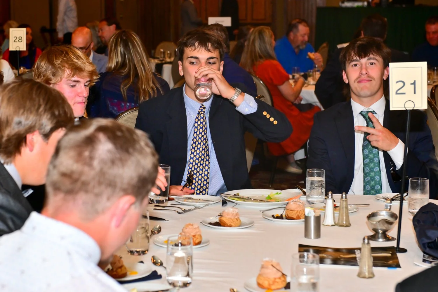 Young men in formal attire sitting at a banquet table, one drinking water and the other making a peace sign, during a formal event or dinner.