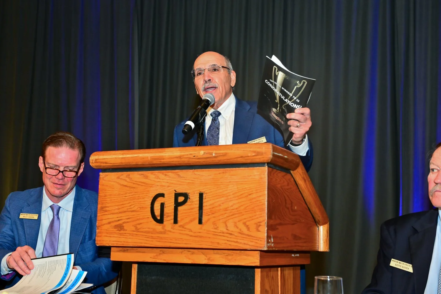 Man in a blue suit speaking at a podium with 'GPI' on it, holding a paper, with two seated men in suits listening, one reading a brochure, all on a stage with dark curtains.