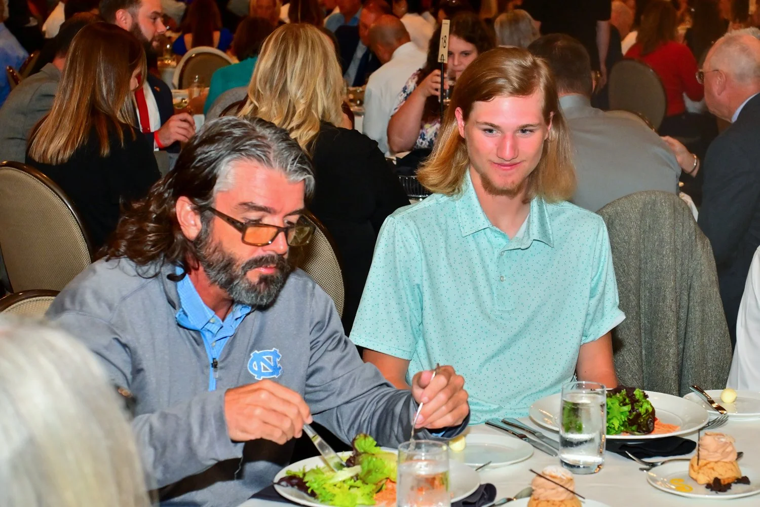 Two men sitting at a formal dining event, enjoying a meal with salad and drinks. The man on the left has long gray hair, beard, glasses, and is wearing a gray jacket with a blue logo. The man on the right has shoulder-length blond hair and a light bl