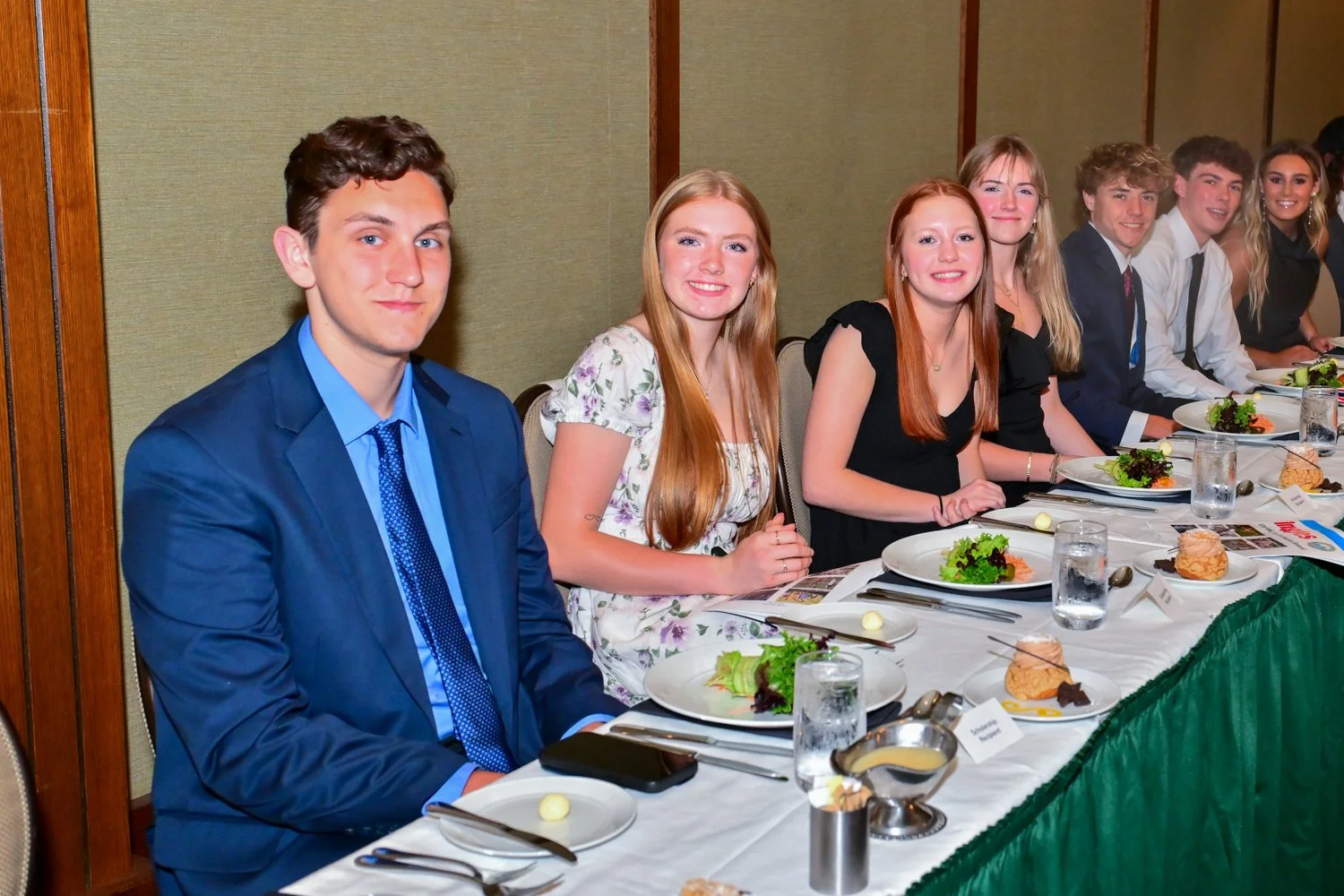 Group of young men and women in formal attire sitting at a dinner table during an event, with plates of food and drinks in front of them.