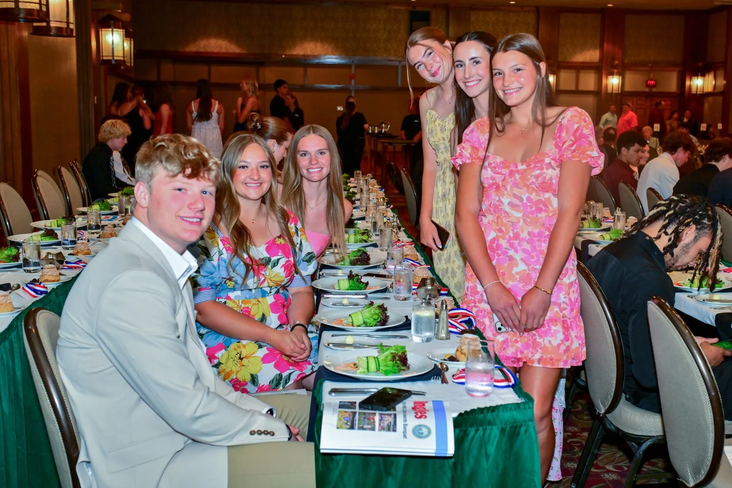Group of six young people, four women and two men, dressed in colorful attire, sitting and standing around a banquet table at a formal indoor event with other guests in the background.