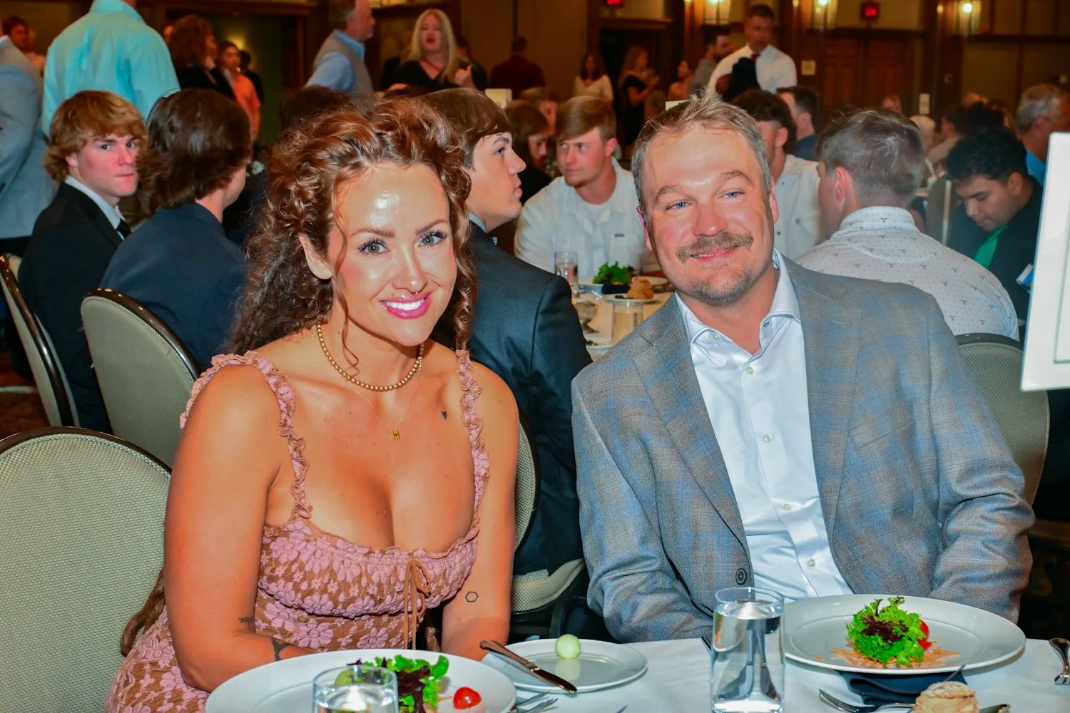 A smiling woman with curly brown hair and light skin, wearing a pink dress with ruffled straps, sits at a table with a man with light skin and short gray hair, wearing a gray plaid blazer and white shirt, at a formal dinner event.