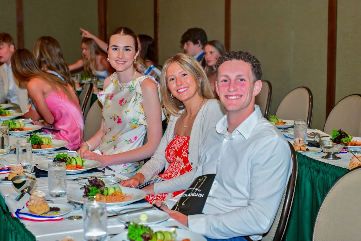 Group of young adults sitting at a formal banquet table, smiling, with plates of food and drinks, in a decorated event hall.