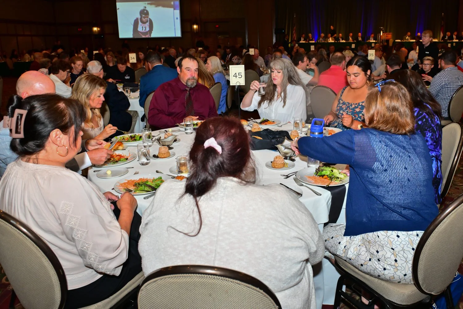 People seated at a round banquet table enjoying a meal during an event with a large crowd, a stage, and a big screen in the background.