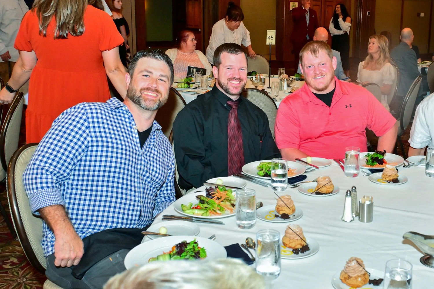 Three men sitting at a banquet table with salads and desserts, smiling at the camera, in a formal event setting with other guests in the background.