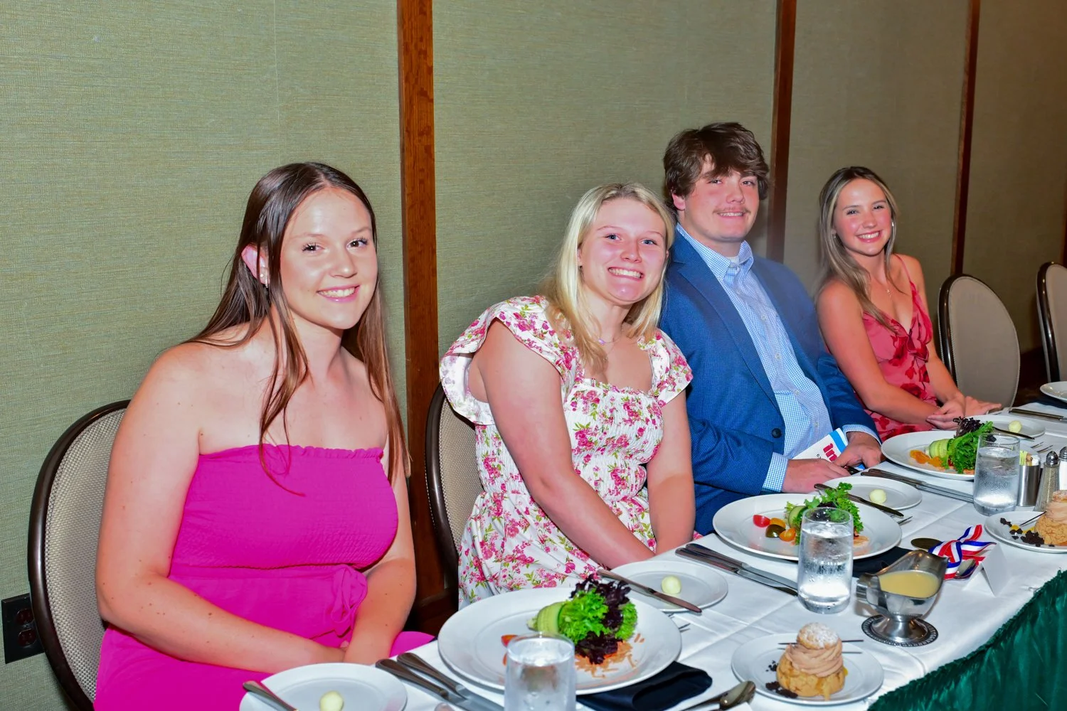 Four young adults sitting at a banquet table with plated salads, water glasses, and desserts, dressed in semi-formal attire for a celebration or event.