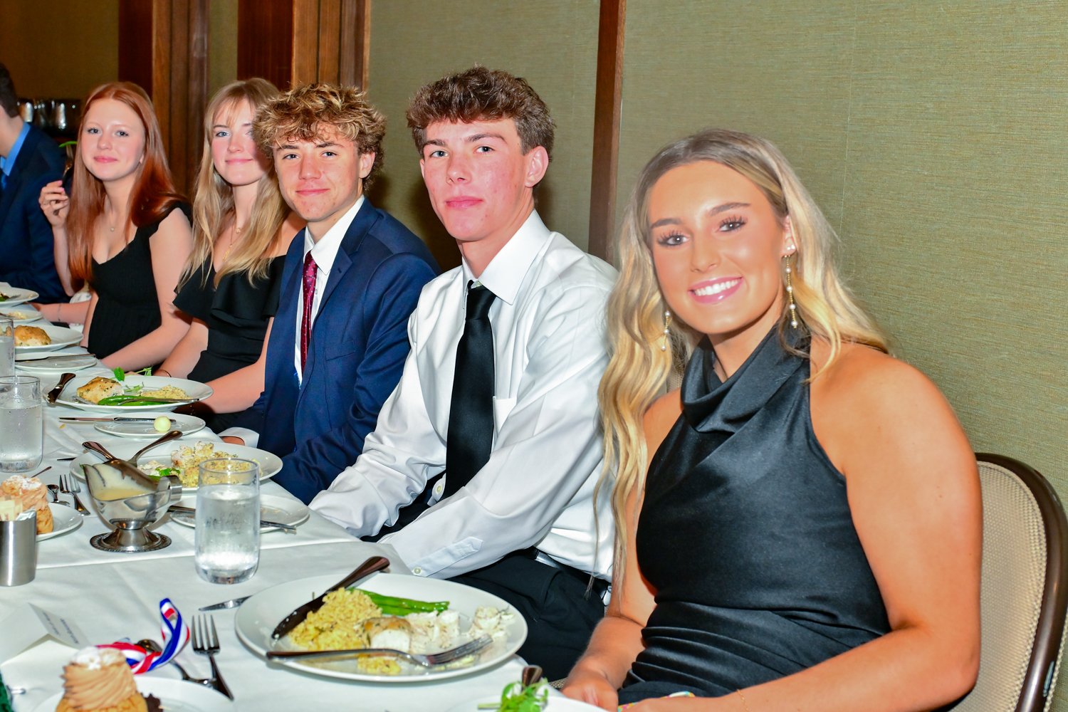 People sitting at a dinner table during a formal event, with plates of food and glasses of water in front of them.