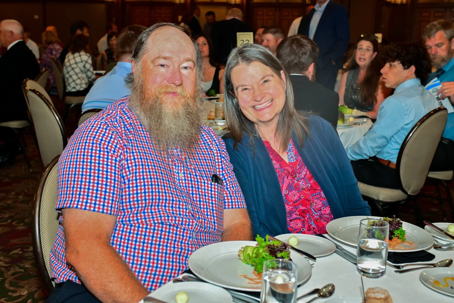 A man with a long beard and a woman smiling at a formal dinner event, sitting at a table with plates of salad, glasses of water, cutlery, and a napkin.
