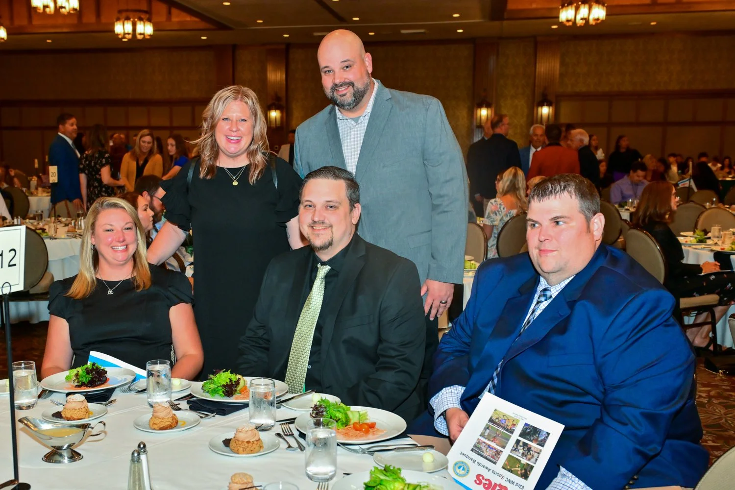 A group of five people at a formal event, three seated at a table and two standing behind them, in a banquet hall filled with other guests and decorated tables.
