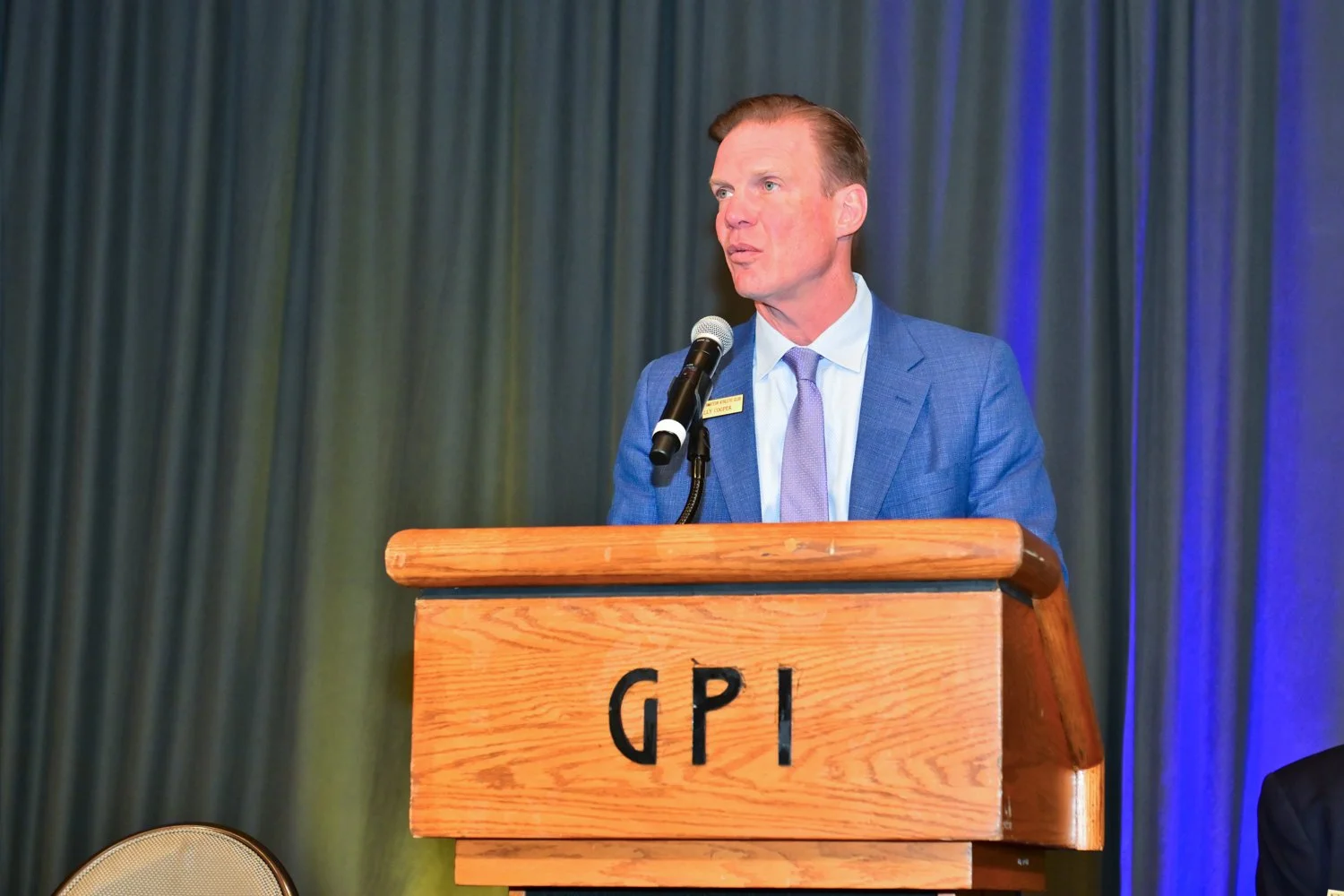 Man in blue suit and lavender tie giving a speech at a wooden podium labeled GPI, with dark curtains in the background.