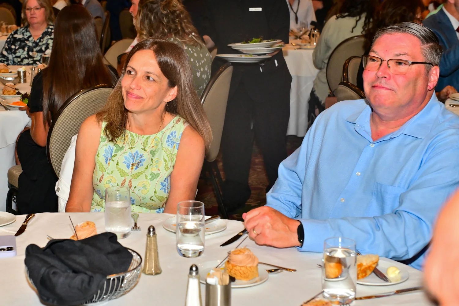 A woman in a floral sleeveless dress and a man in a light blue shirt sit at a dinner table during a formal event, with plates of cake, glasses of water, and silverware visible on the table. Other guests are seated in the background.