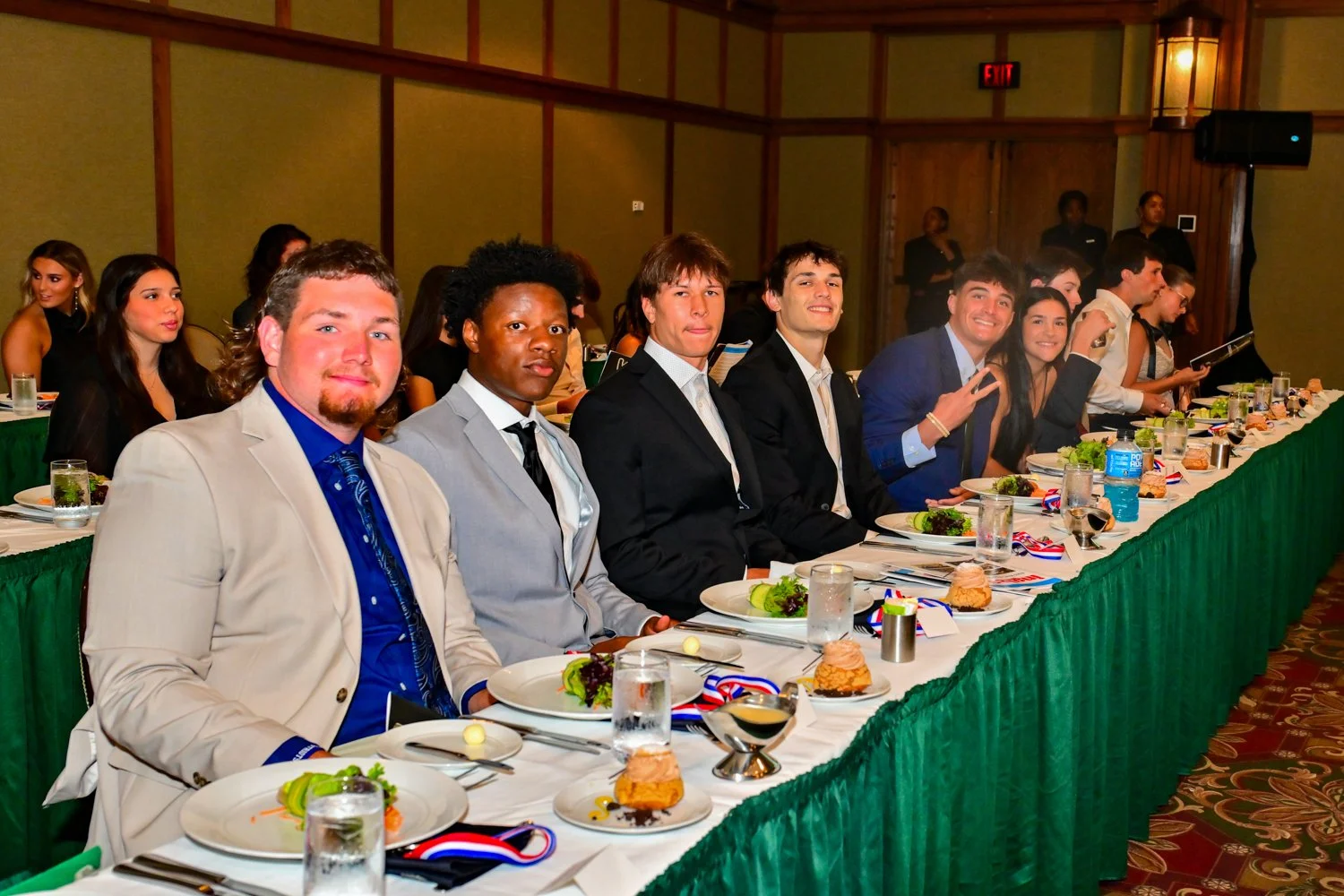 Group of young adults sitting at a banquet table, dressed in formal attire, smiling and making peace signs during a formal event or celebration.