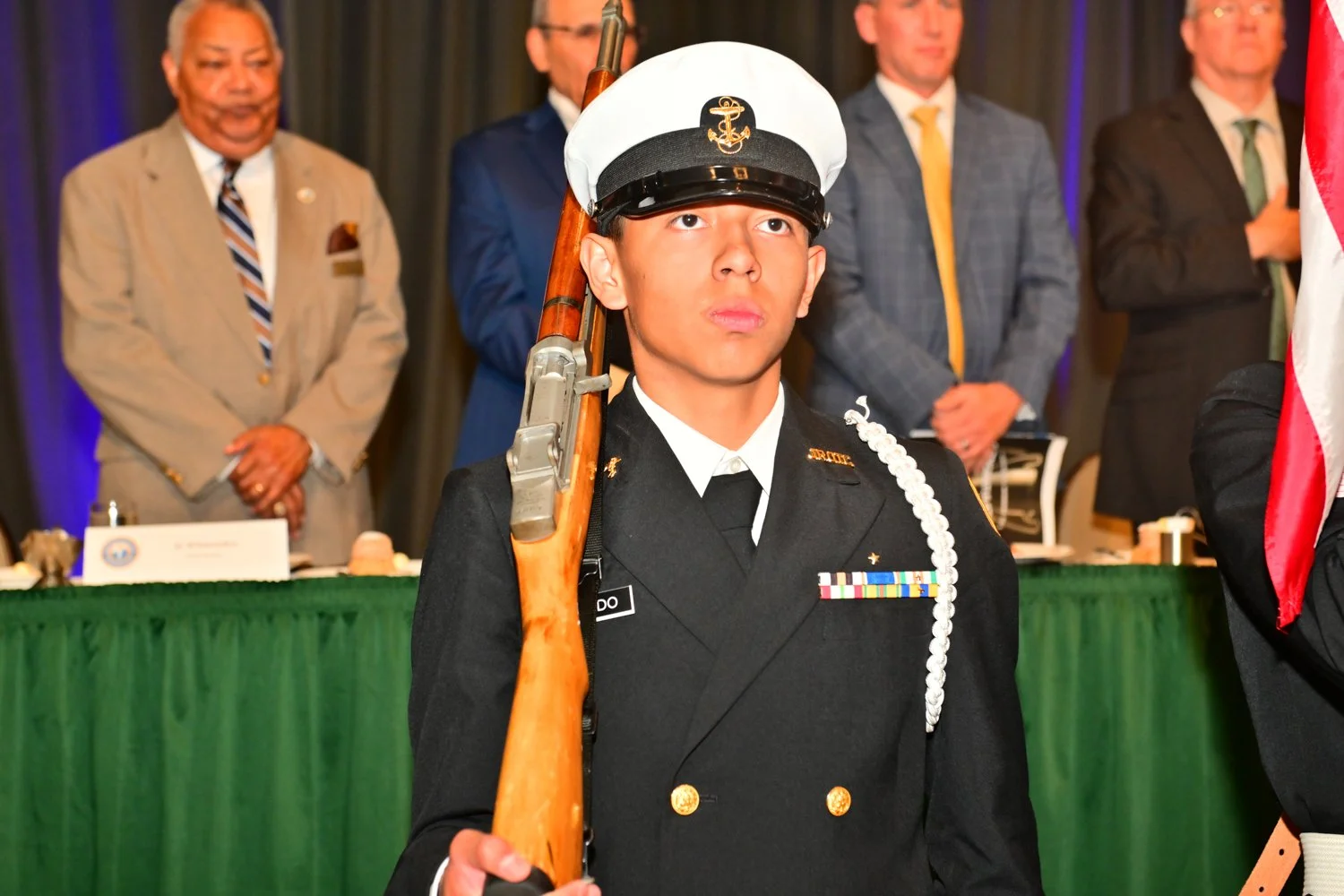 A young man in a military dress uniform holding a rifle, standing in front of a green table with officials and onlookers behind him at a formal ceremony.