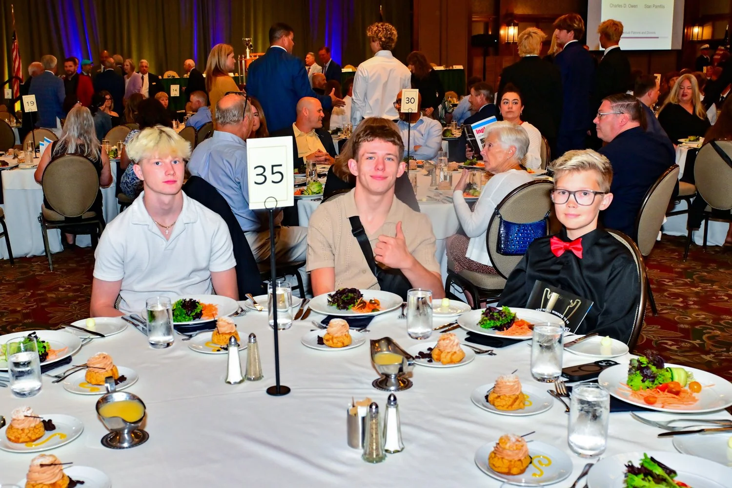A banquet hall filled with people at round tables, with three young boys sitting at one table in the foreground. The boy on the left has blonde hair, is wearing a white shirt, and is looking at the camera. The boy in the middle is giving a thumbs-up,