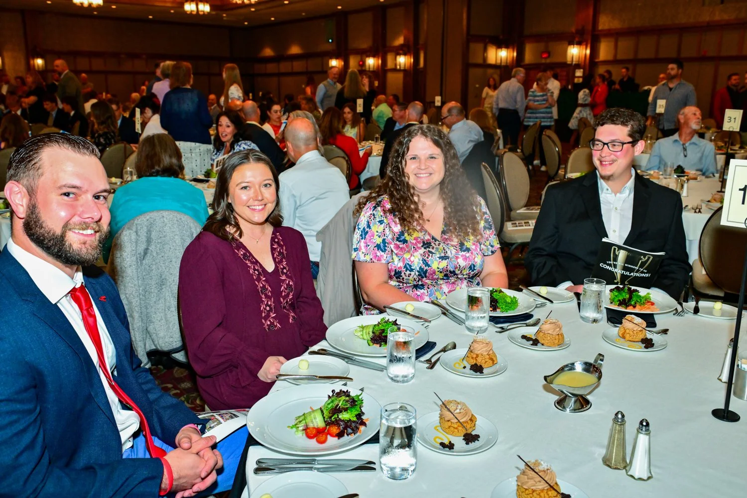 Four people sitting at a round banquet table with plates of salad and dessert, at a formal event or gathering in a large, decorated hall with many other guests seated at similar tables in the background.