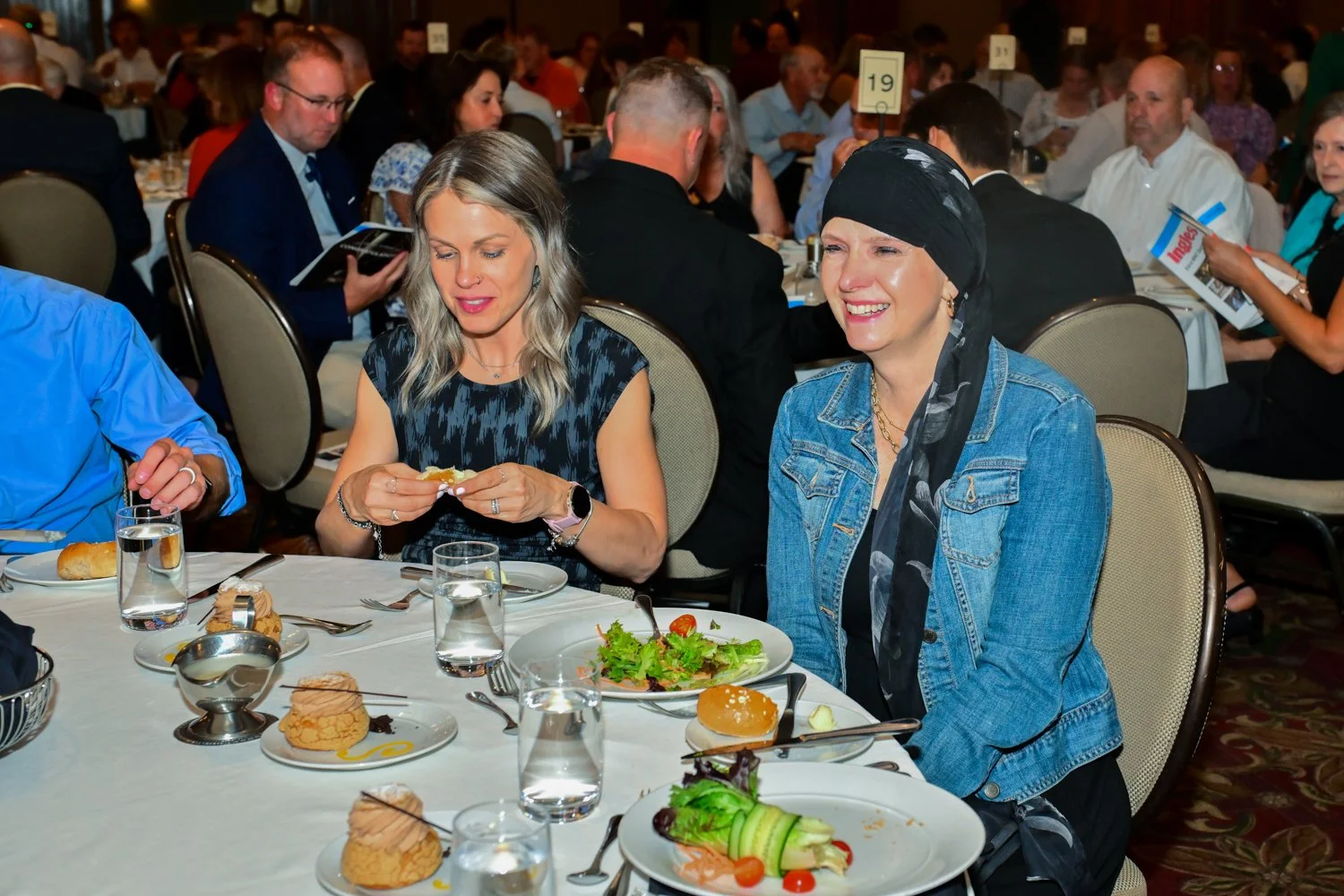 Two women sitting at a banquet table during a formal event, enjoying their meal amidst many other attendees in the background.