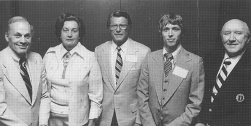 Black and white photo of five individuals in vintage suits posing for a photo in front of a dark background.
