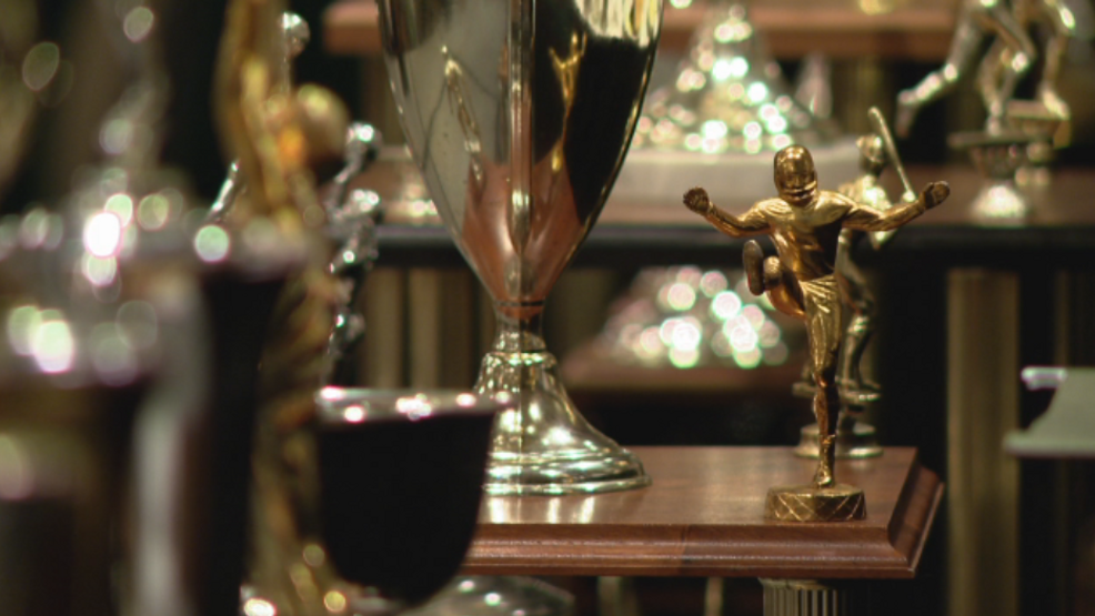 A football player trophy, a large silver trophy, and other sports trophies displayed on a wooden shelf.