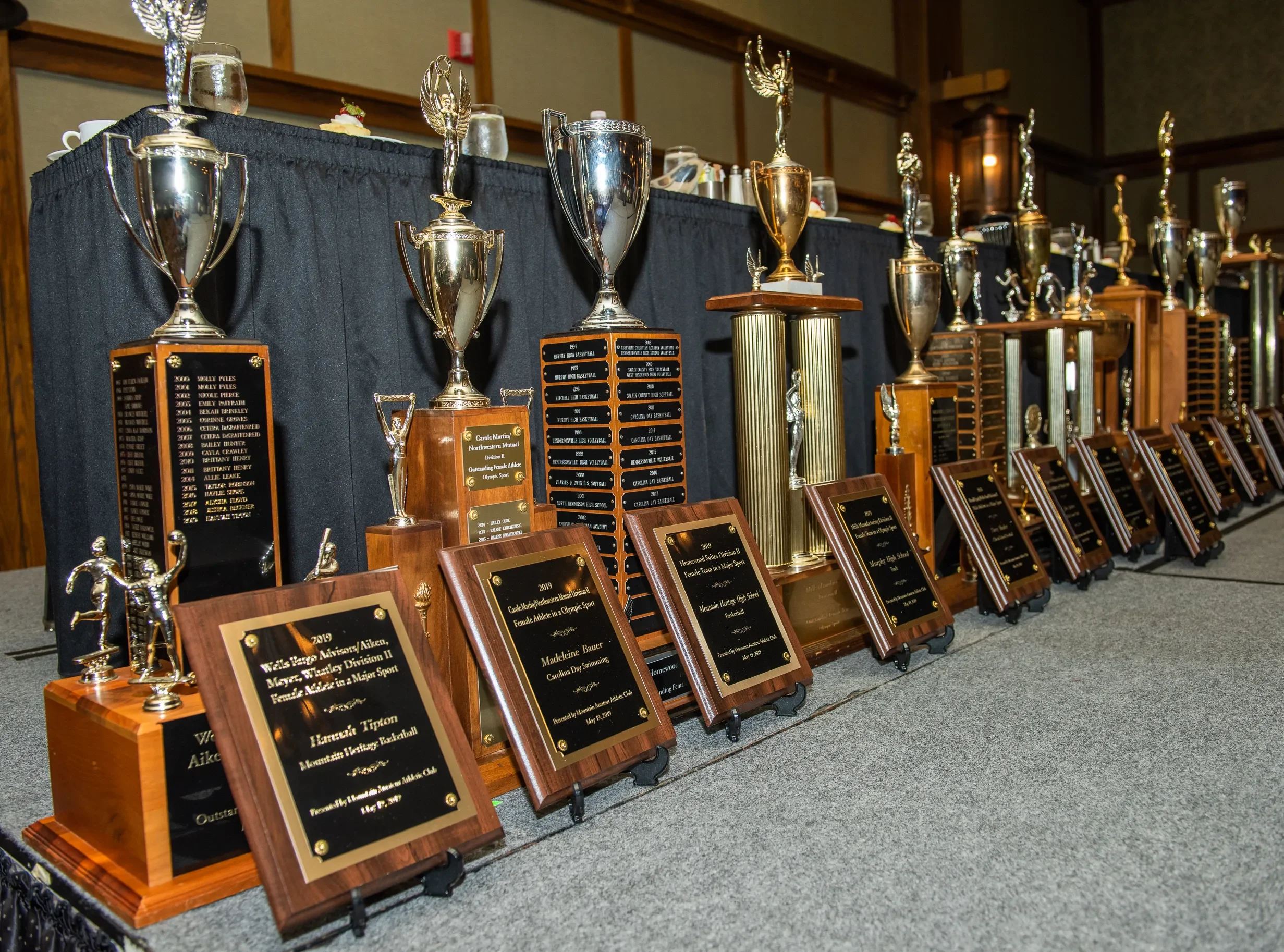 A display of various trophies and awards on a table, with plaques and smaller trophies in the foreground and larger trophies in the background, some with figures of athletes on top, set in an indoor event space.