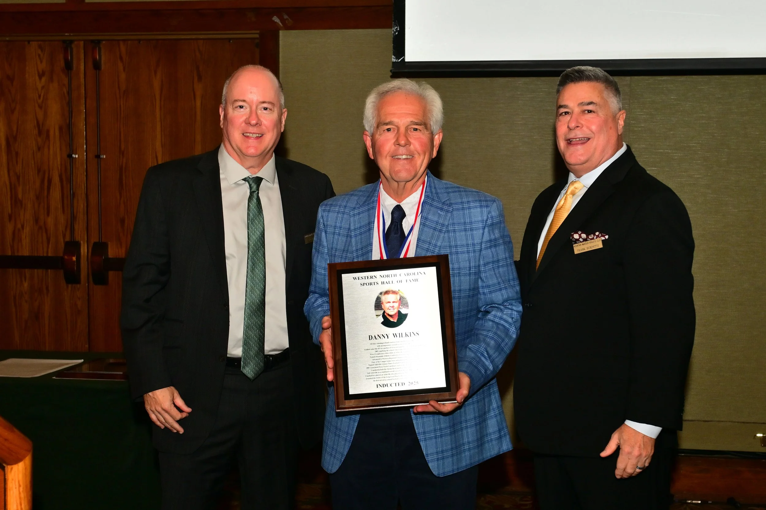 Three men in suits smiling at a ceremony, one man in the middle holding a framed certificate that reads 'Western North Carolina Sports Hall of Fame, Danny Wilkins, Inducted 2023'.