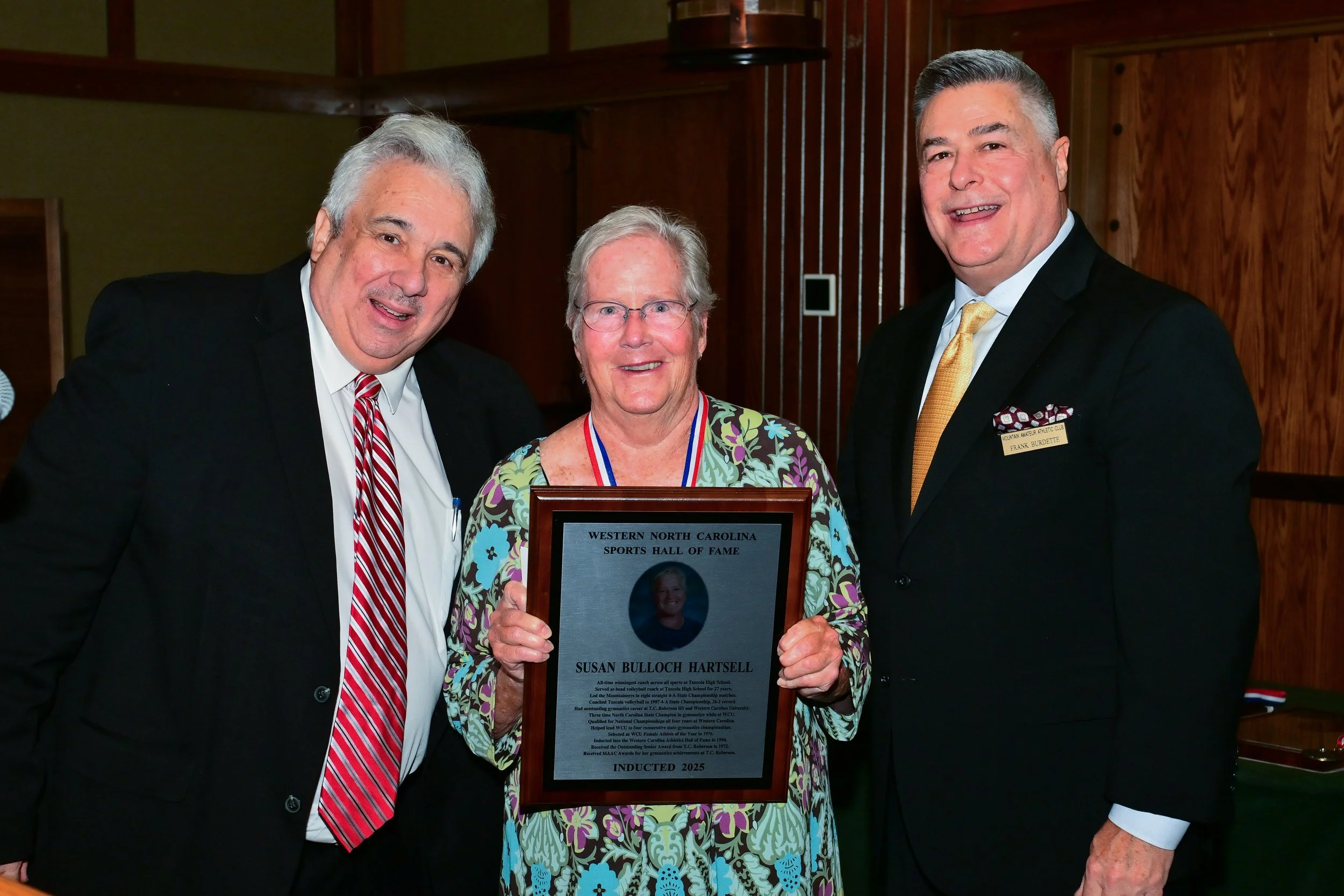 Three people standing together indoors. The woman in the center is holding a framed plaque that says 'Western North Carolina Sports Hall of Fame' and 'Inducted 2025.' The man on the left wears a black suit with a white shirt and red striped tie, and the man on the right wears a dark suit with a white shirt and yellow tie. The woman is wearing glasses and a colorful floral blouse. They are all smiling.
