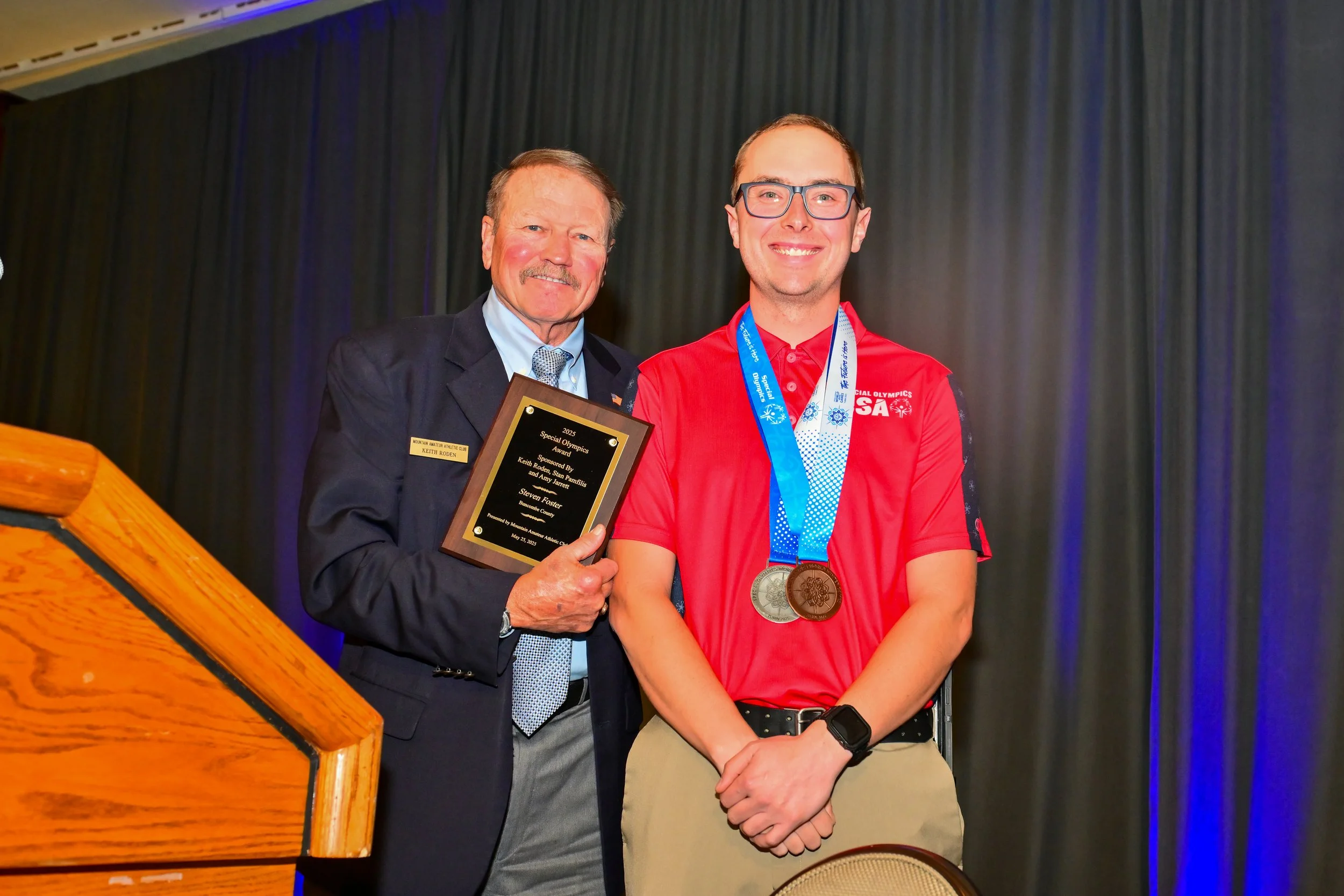 An older man in a suit and tie presenting an award plaque to a young man wearing glasses, a red polo shirt, and medals around his neck at an indoor event.