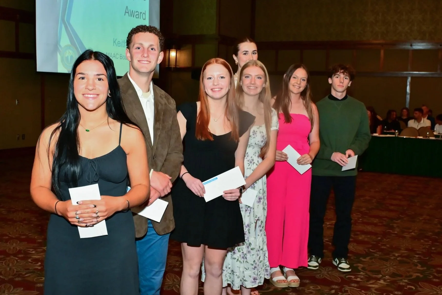 Group of seven young adults at an awards ceremony, holding certificates, standing in a line inside a decorated banquet hall.
