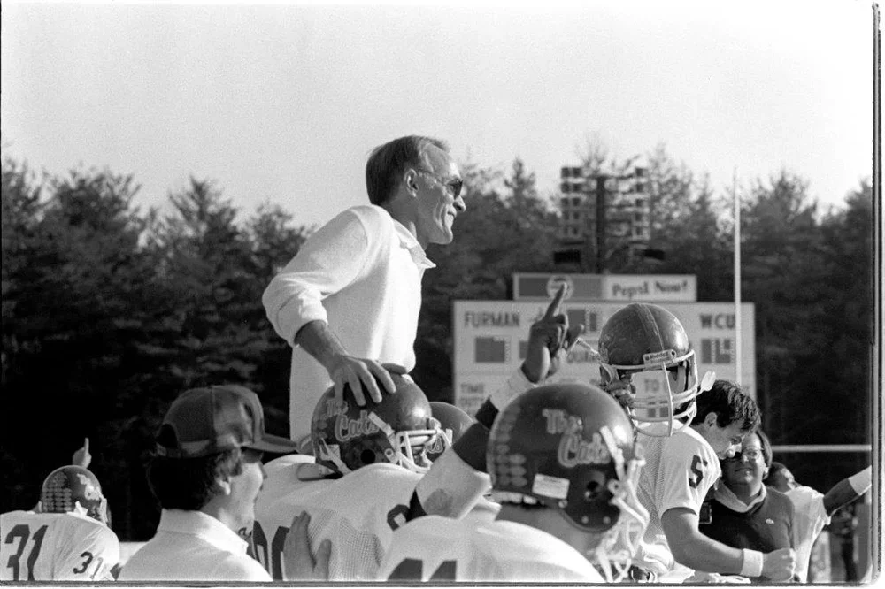 A football coach is speaking to a team of young football players on the field, wearing helmets and uniforms, with a scoreboard in the background.