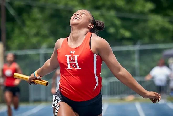 A female athlete in a red sleeveless running uniform celebrating after a race, holding a relay baton in one hand, smiling with her head tilted back, on an outdoor track.