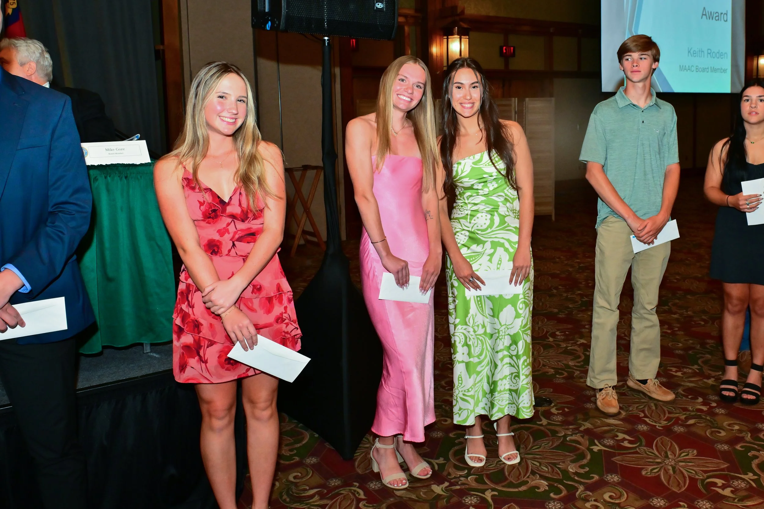 Group of young women and men standing in a line at an indoor event, smiling for the camera, holding papers or envelopes, dressed in colorful and stylish summer clothing.
