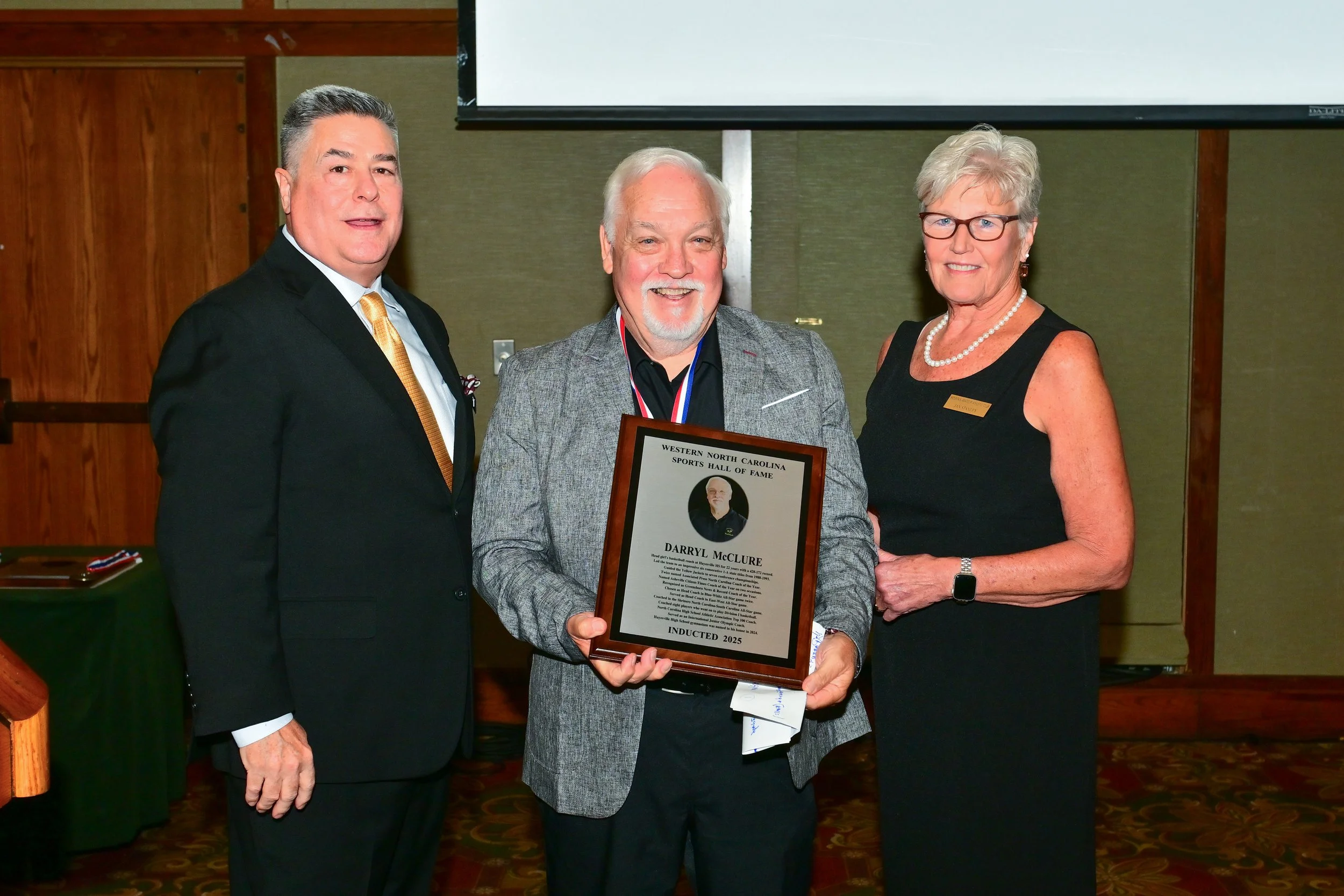 A group of three people standing indoors, with two men and one woman. The man in the middle is holding a framed certificate or award, smiling. The woman on the right has short gray hair, wears glasses, a black sleeveless top, a pearl necklace, and a watch. The man on the left has dark hair with gray, is dressed in a black suit with a white shirt and a gold tie.