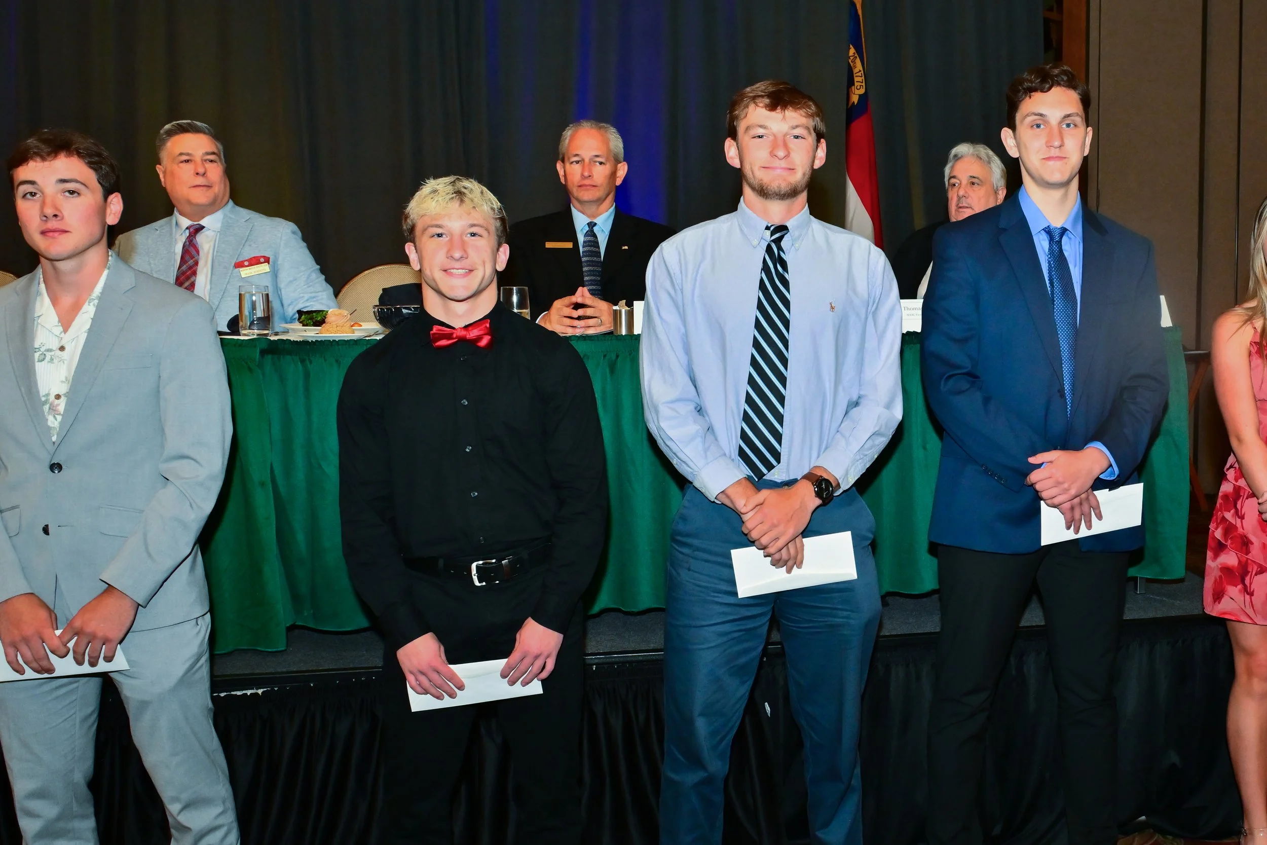 Young men dressed in formal attire, standing in front of a panel at an indoor event. Some are holding papers, with a backdrop featuring a flag and dark curtains.