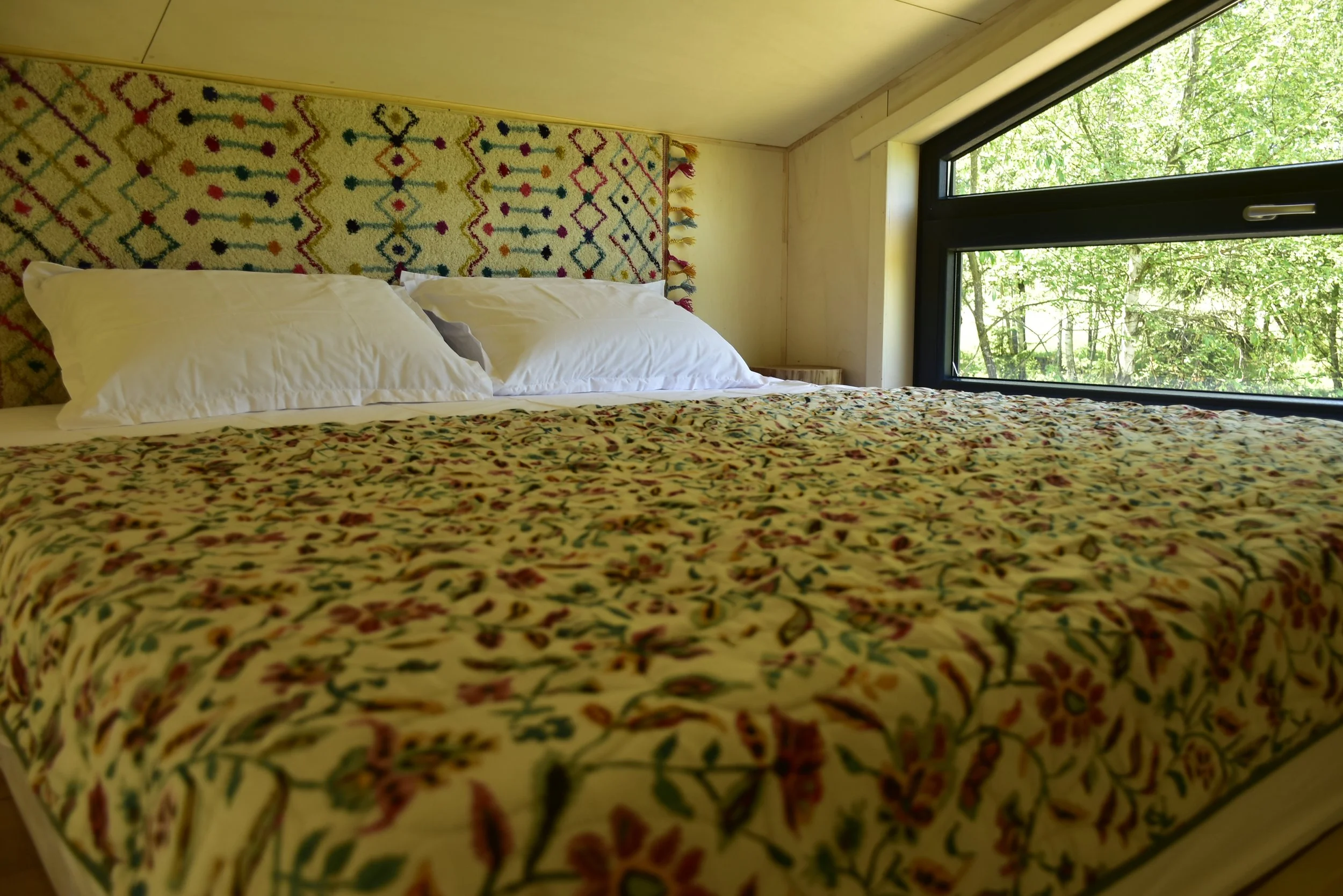 WildKind Nature Retreat Bucovina Romania Tara Dornelor - Close-up view of a loft bed with colorful patterned bedding and white pillows, against a wall with a decorative headboard, next to a large window with a green outdoor view.