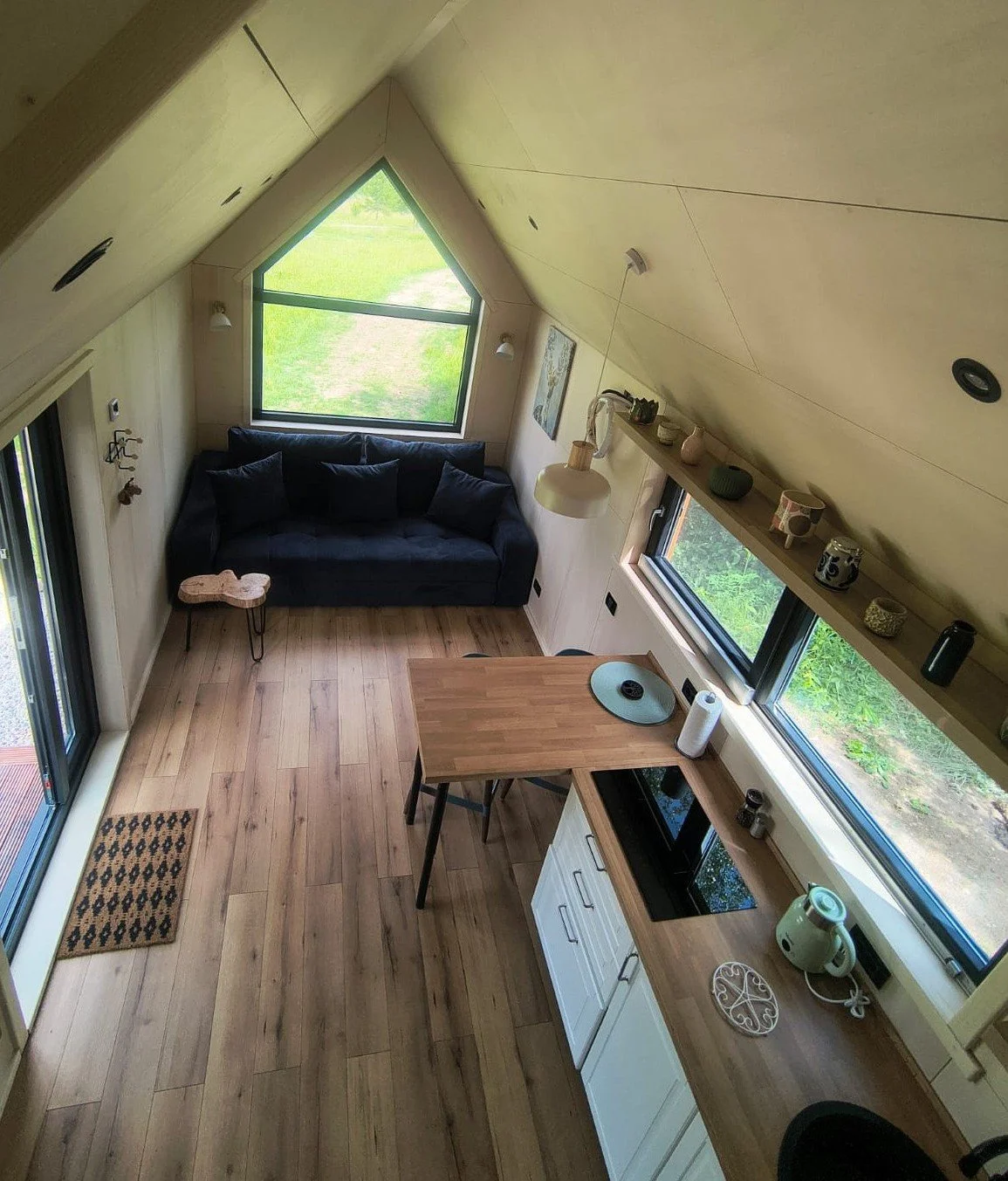 WildKind Nature Retreat Romania Interior view of a cozy tiny house, with a small kitchen, decorative items on a shelf, a deep blue sofa, and a small coffee table. The space has wooden floors sloped ceiling, and large windows letting in natural light.