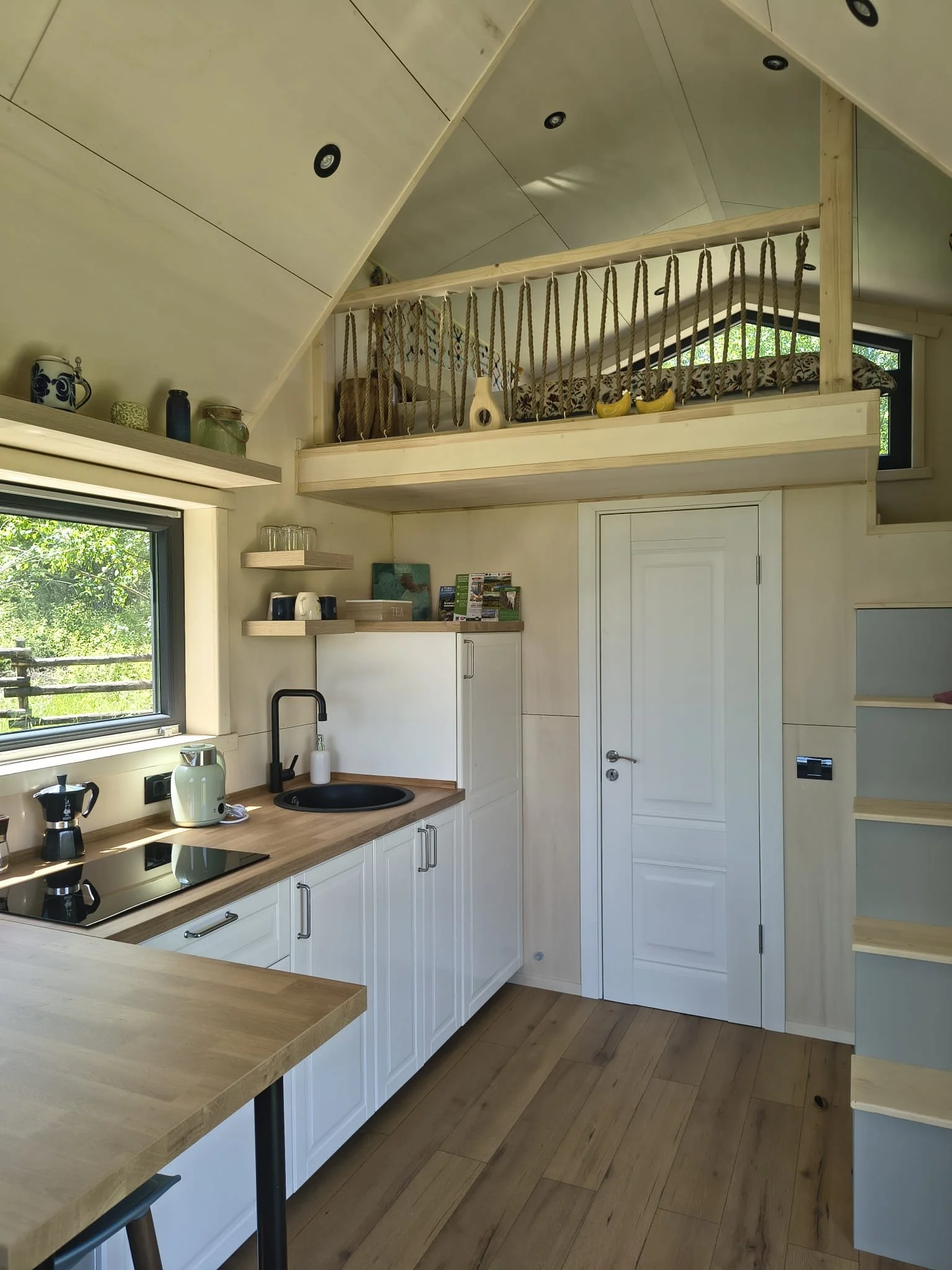 Wildkind Nature Retreat Tara Dornelor Interior of a tiny house modern kitchen with a wooden countertop, white cabinets, black sink, and a window showing greenery outside. There is a small loft area with a wooden railing and knotted ropes for support.