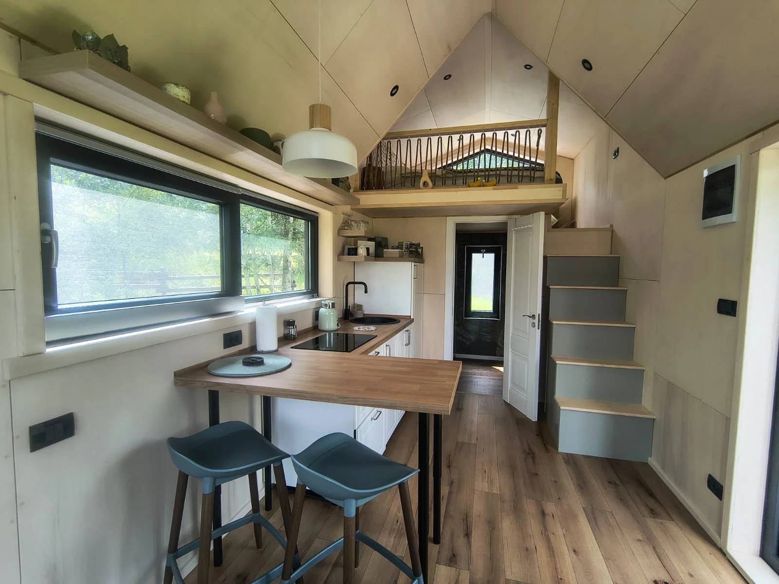 WildKind Nature Retreat Tara Dornelor, Bucovina, Romania - Interior of a tiny house with a kitchenette, stairs leading up to a sleeping loft, wooden flooring, and a large window bringing in natural light.