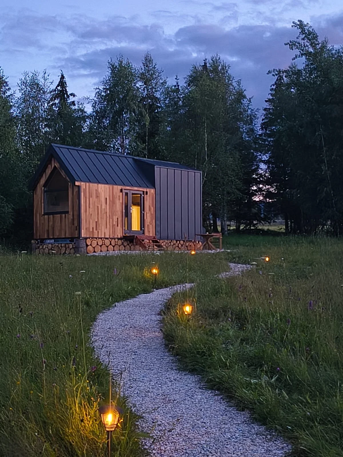 WildKind Nature Retreat Tara Dornelor Bucovina Romania A tiny house with a metal roof is situated in a grassy field at dusk, with a winding gravel path lined by small lanterns leading to the cabin.