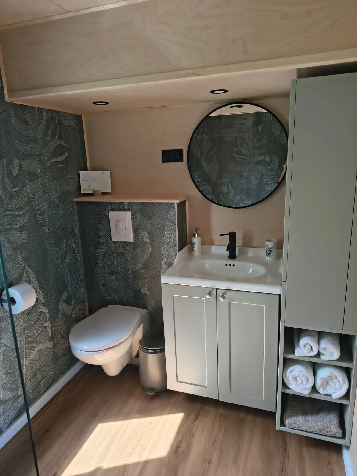 WildKind Nature Retreat Bucovina Tara Dornelor Romania - A modern bathroom with a white toilet, a small white vanity with a sink, a round mirror, open shelves with rolled towels, and patterned wallpaper on one wall.