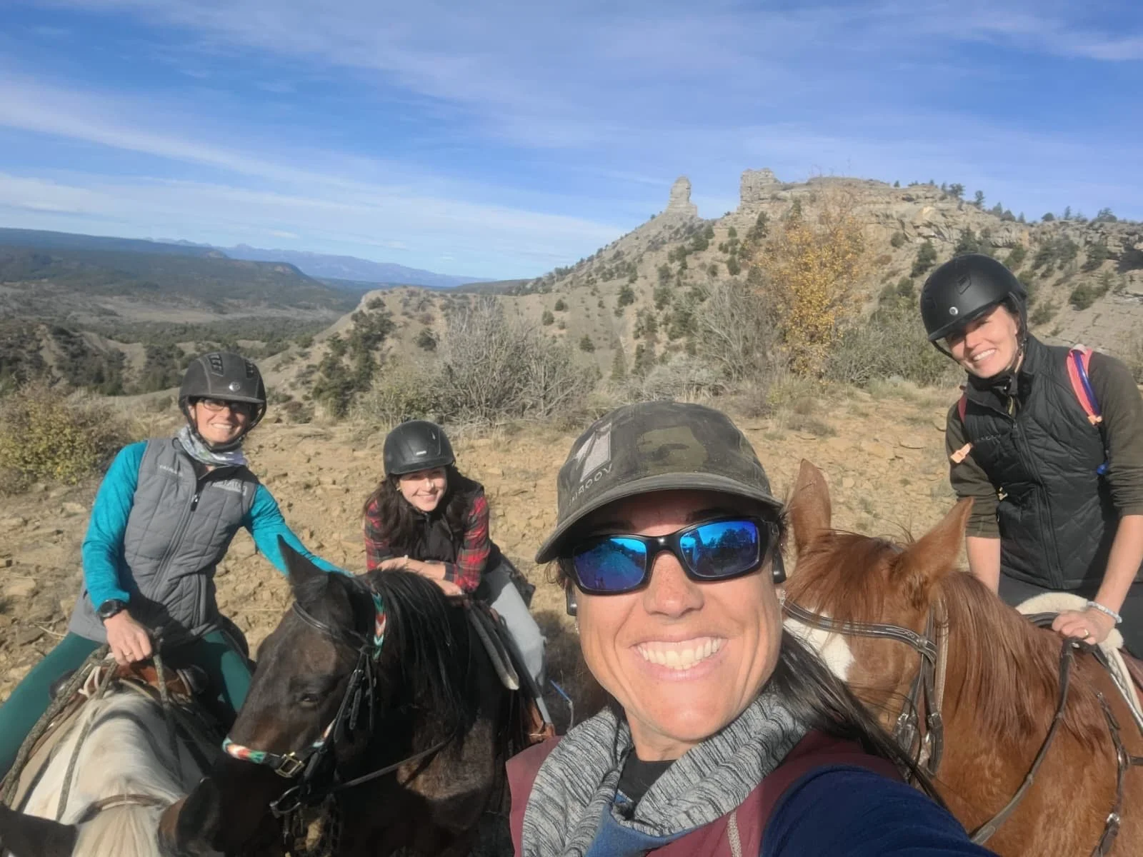 Four women horseback riding in a mountainous landscape with rocky terrain, sparse trees, and a clear blue sky, wearing helmets and outdoor gear.