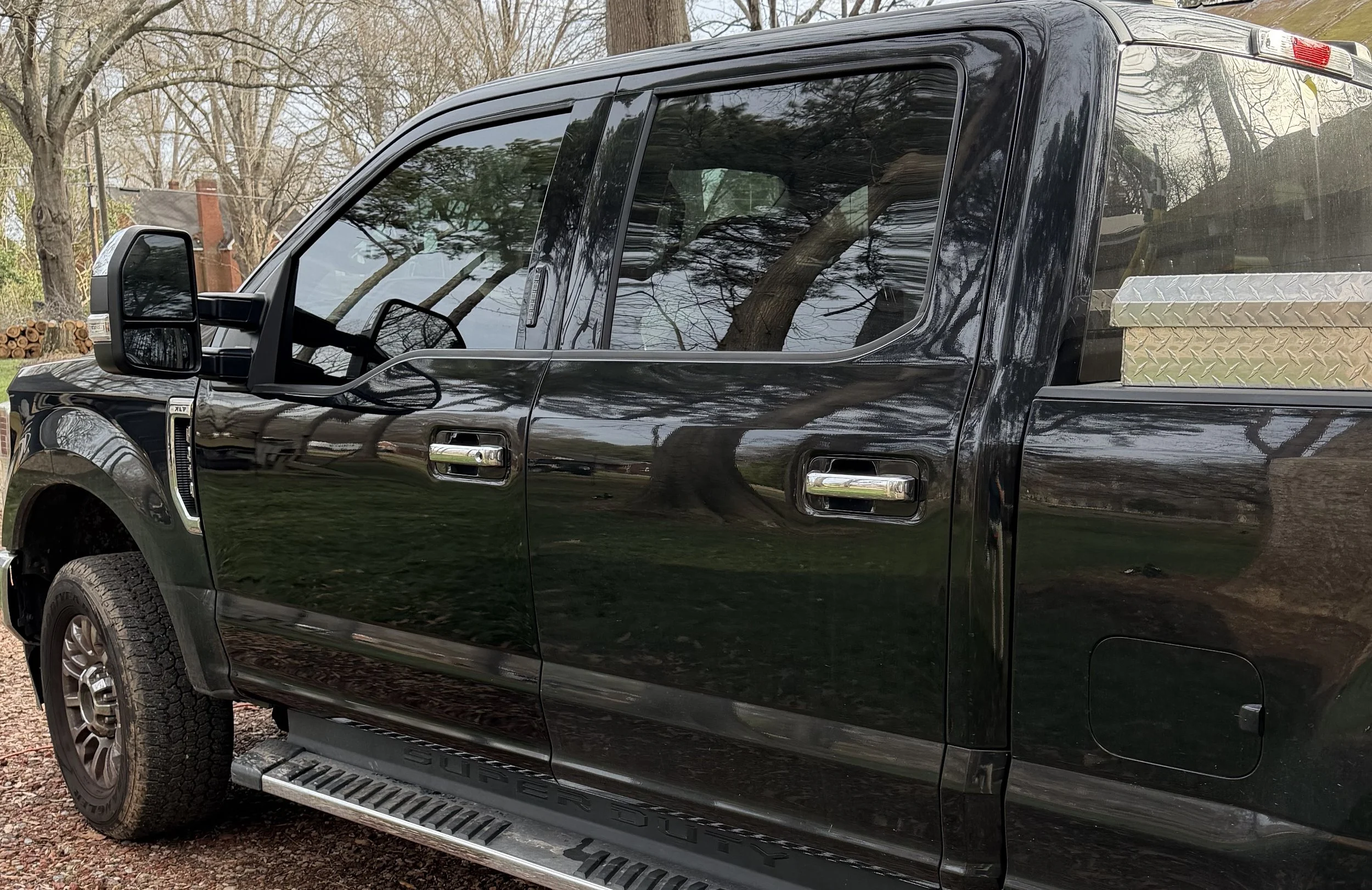 A black pickup truck parked outdoors with trees reflecting on its windows and body, and some logs stacked in the background.