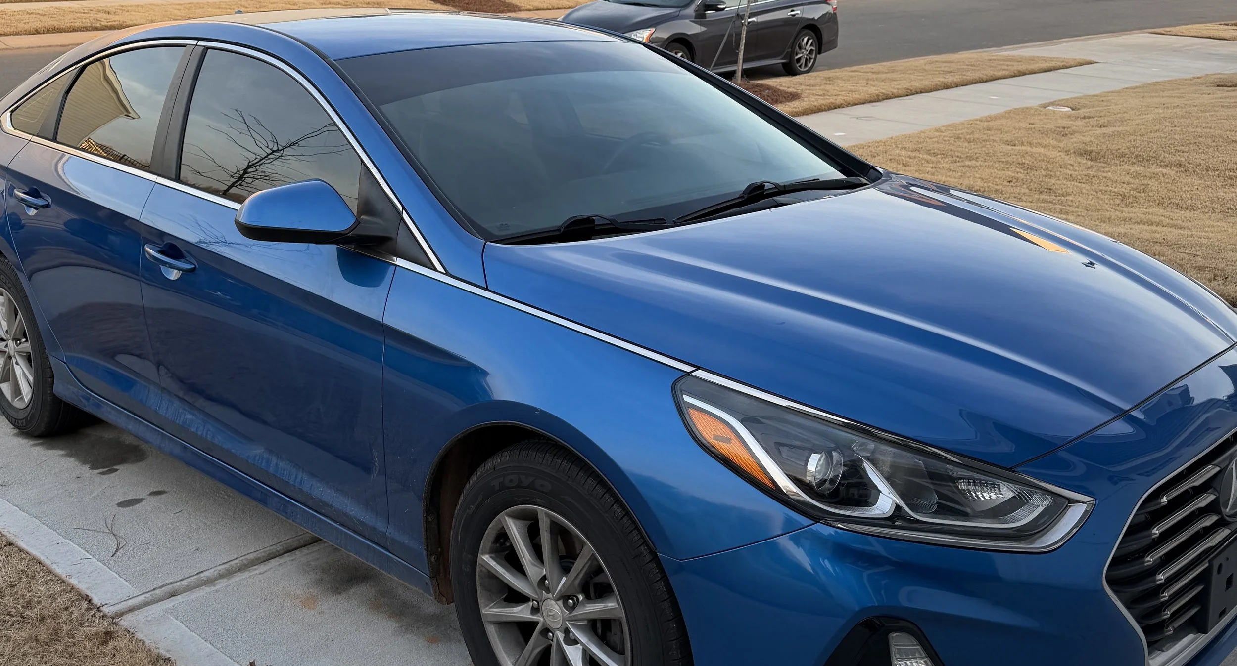 Blue sedan parked on driveway in front of a house with a grassy yard and concrete sidewalk, with a gray car parked on the street in the background.