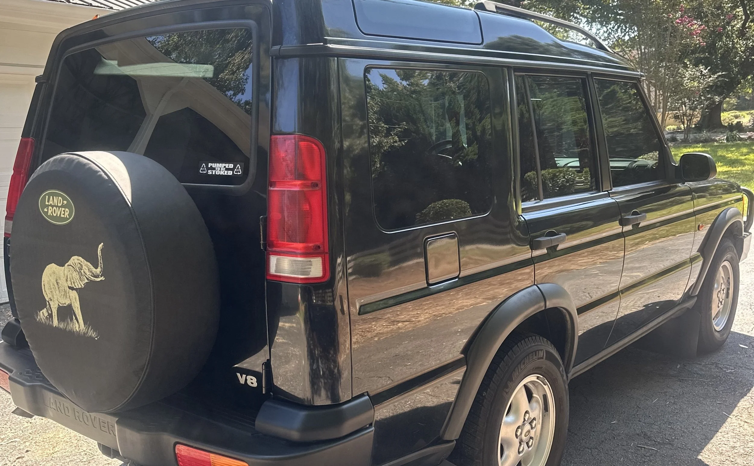 Rear side of a black Land Rover SUV parked outdoors, showing its spare tire cover with an elephant graphic and Land Rover logo, and a window sticker that reads 'Pumped Before Stoked'.