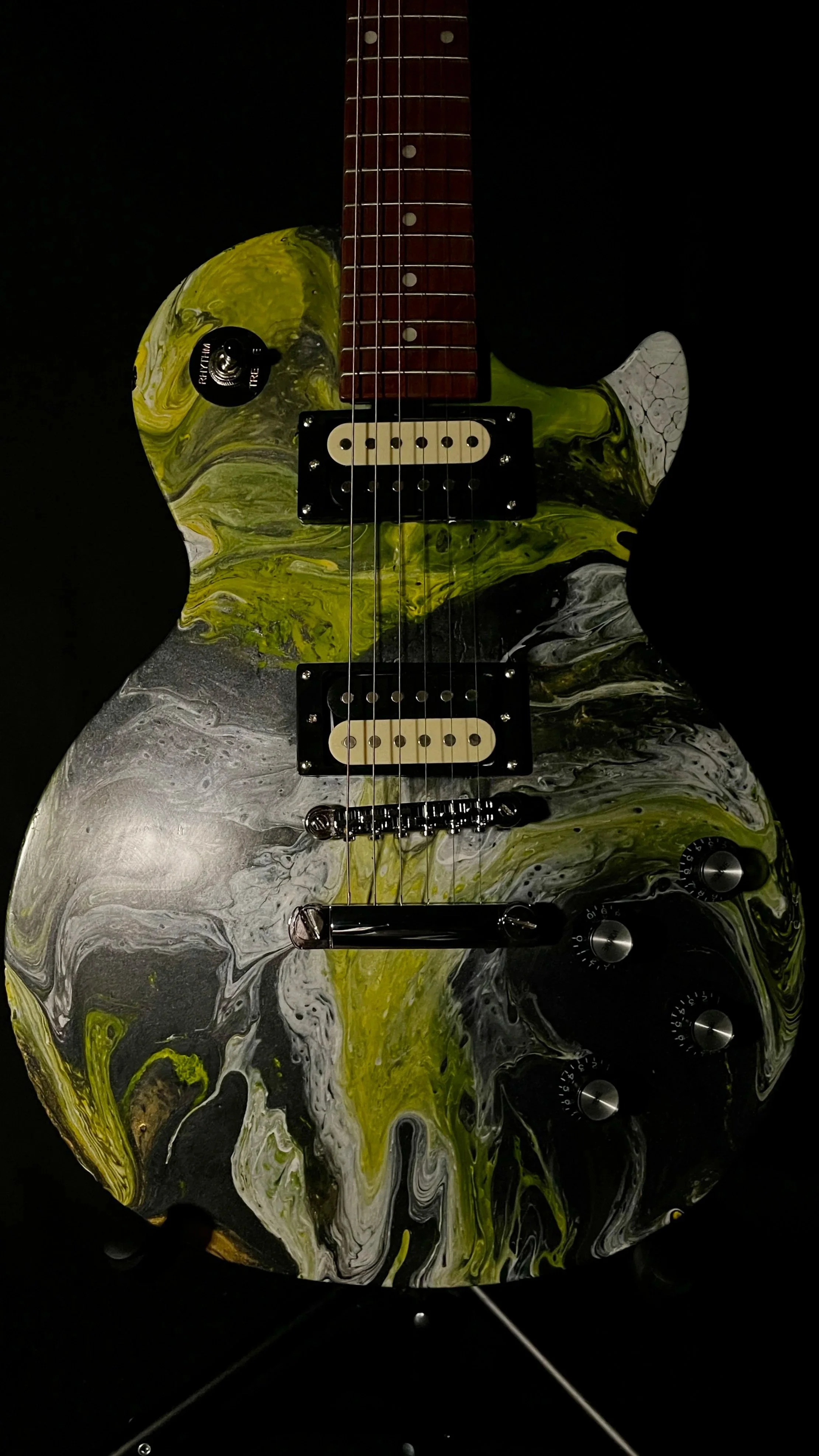 Electric guitar with a marbled black, white, and yellow body, black pickups, and a red-brown neck against a black background.