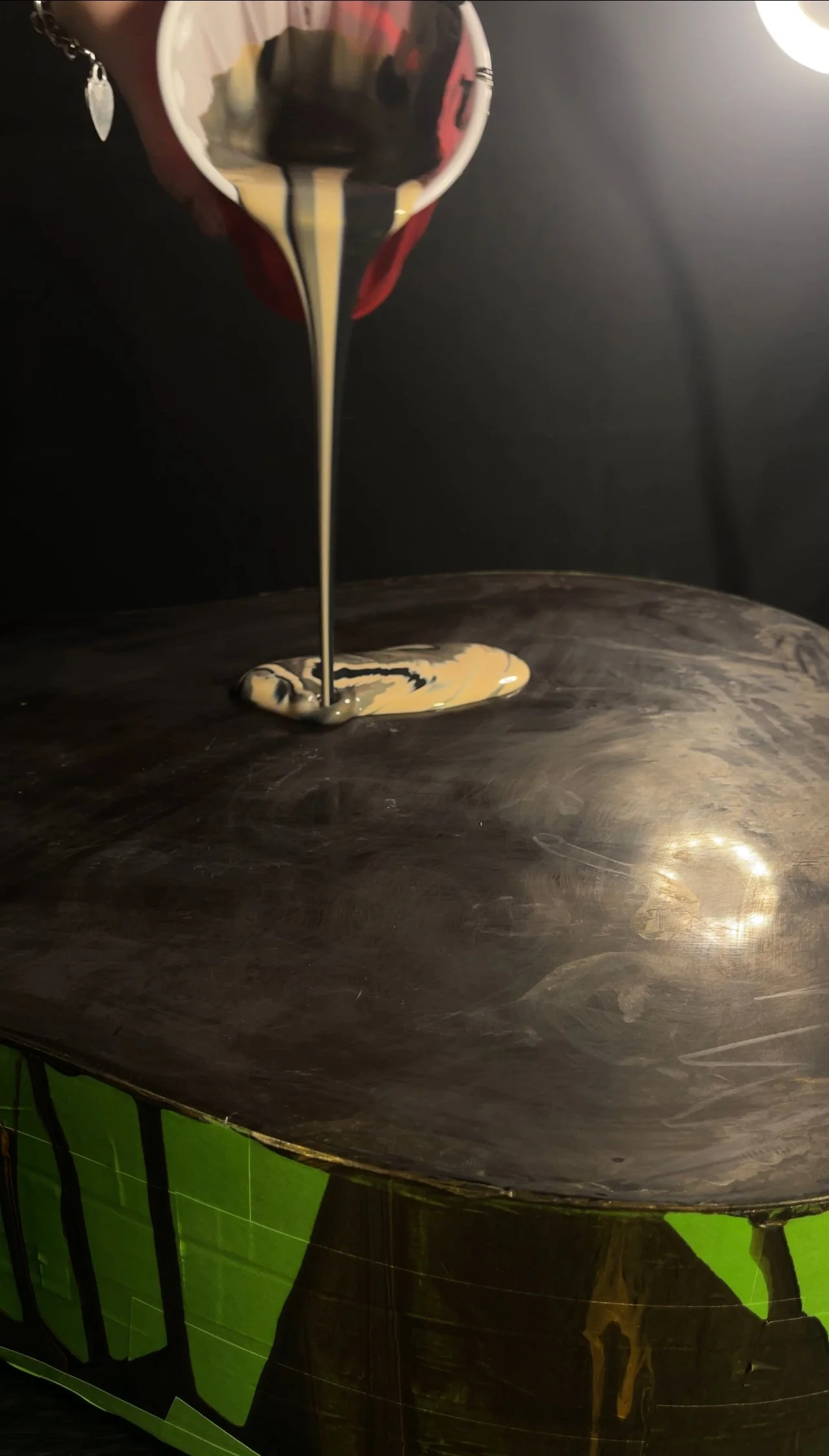 Cream-colored paint is being poured from a cup onto a dark, textured surface.
