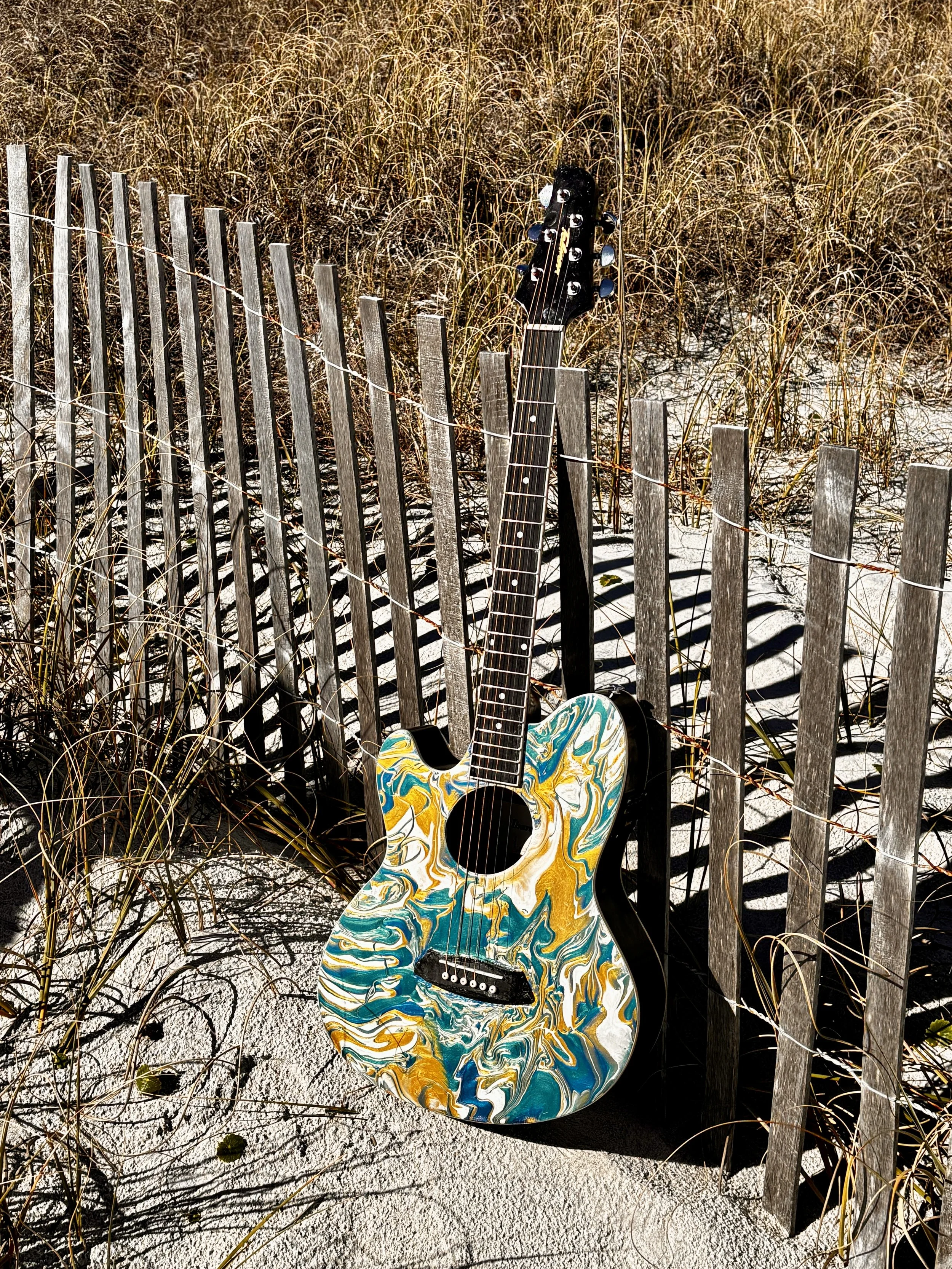An acoustic guitar with a colorful, swirling paint design leans against a weathered wooden fence on a sandy beach, surrounded by tall grass.