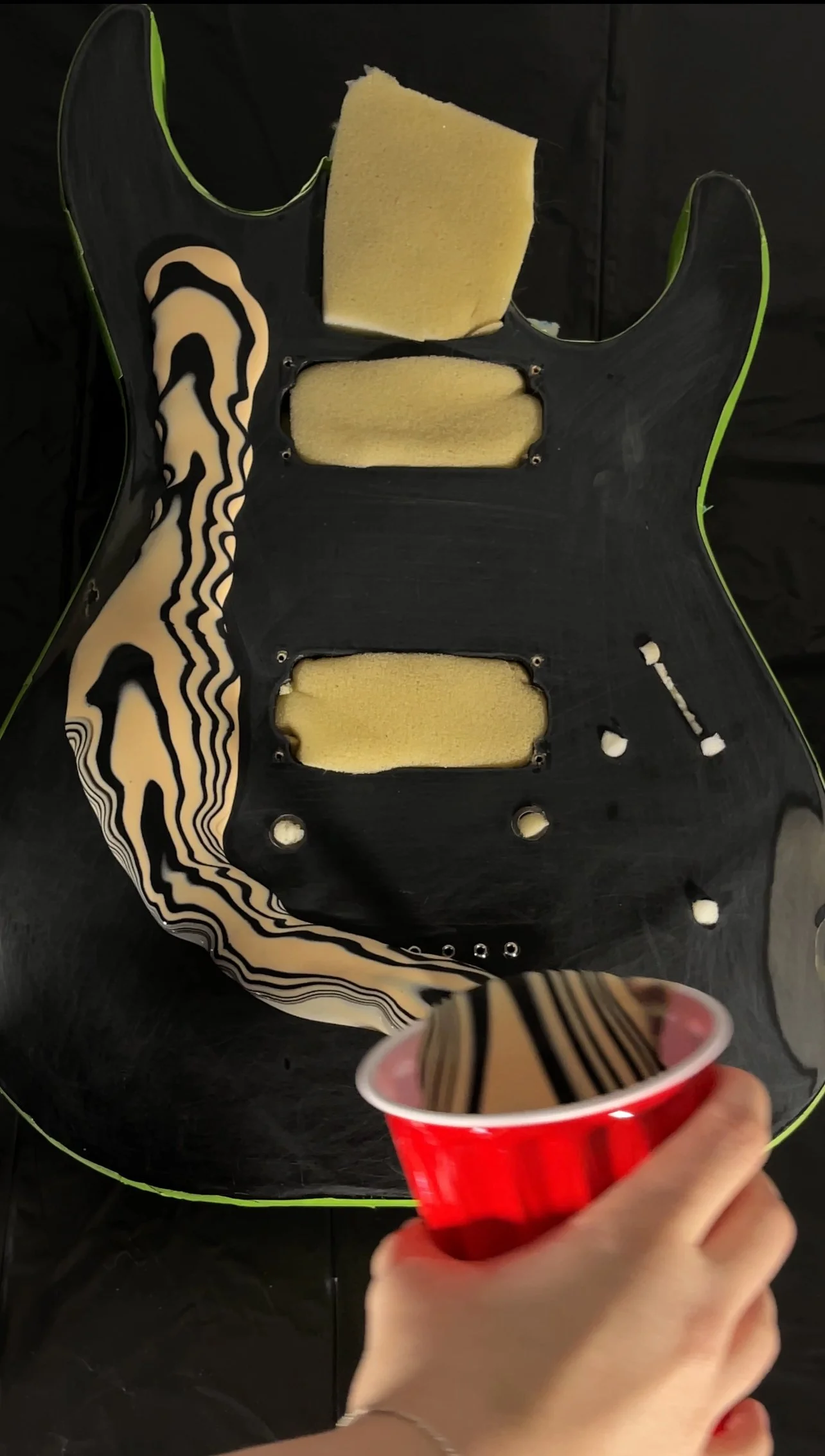 A person holding a red cup with a guitar body in the background. The guitar body has been stripped of its electronic components and paint, revealing foam padding and a black base layer, with a striped black and beige pattern on the left edge.
